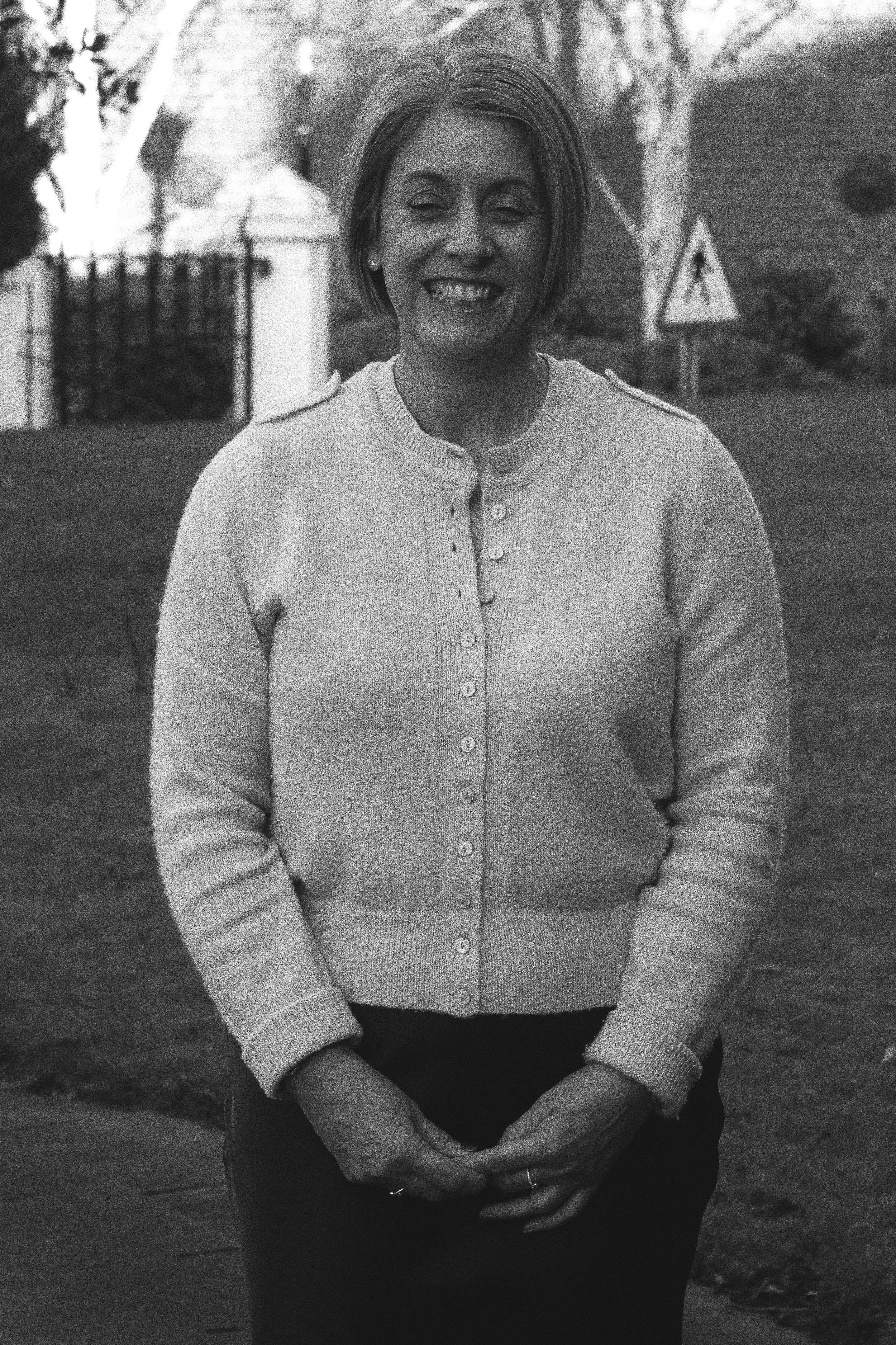 A woman smiling outdoors, wearing a cardigan and standing on a pathway, with trees and a sign in the background.