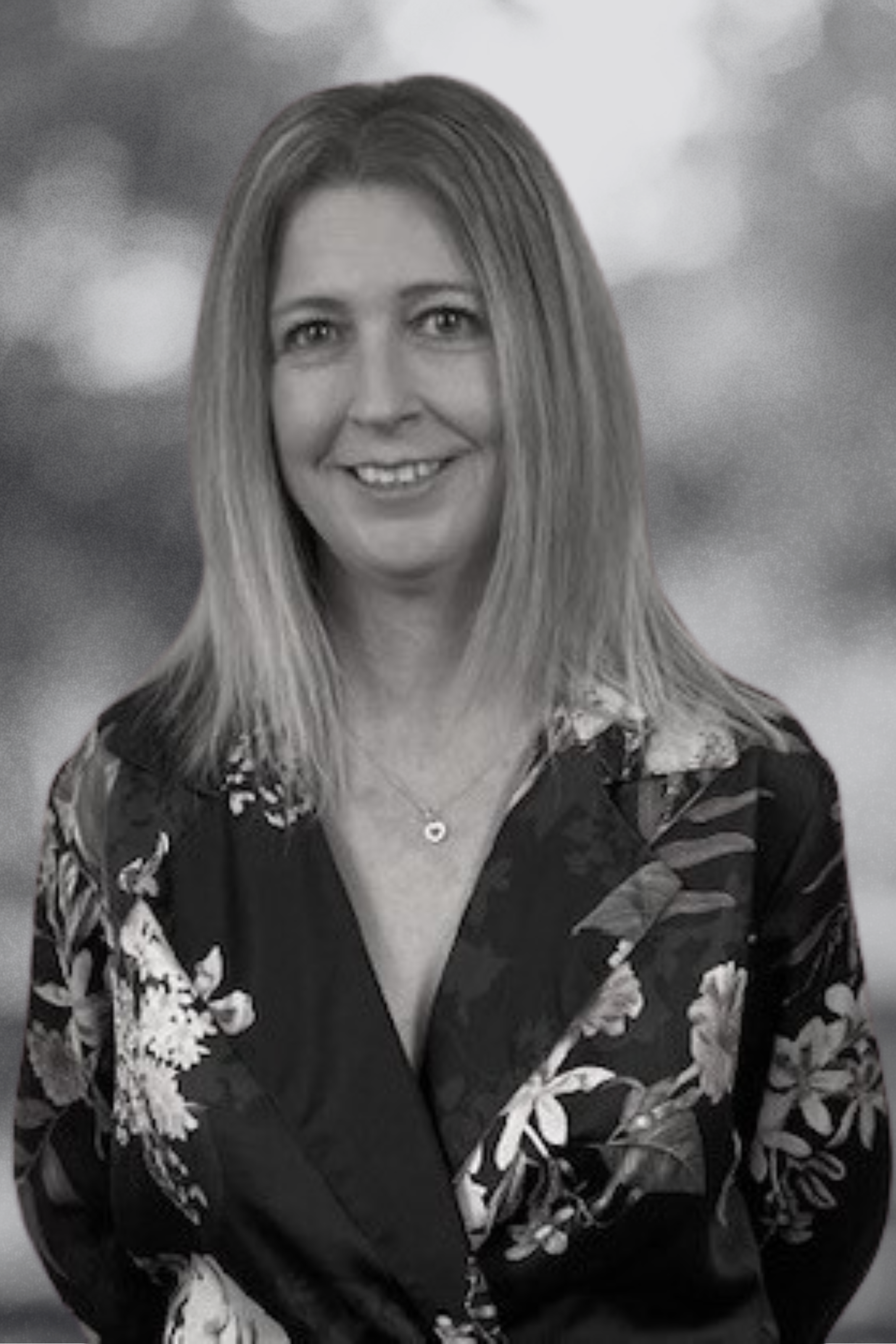 A woman with straight shoulder-length hair smiling at the camera, wearing a floral patterned blouse and a necklace, with a blurred outdoor background.