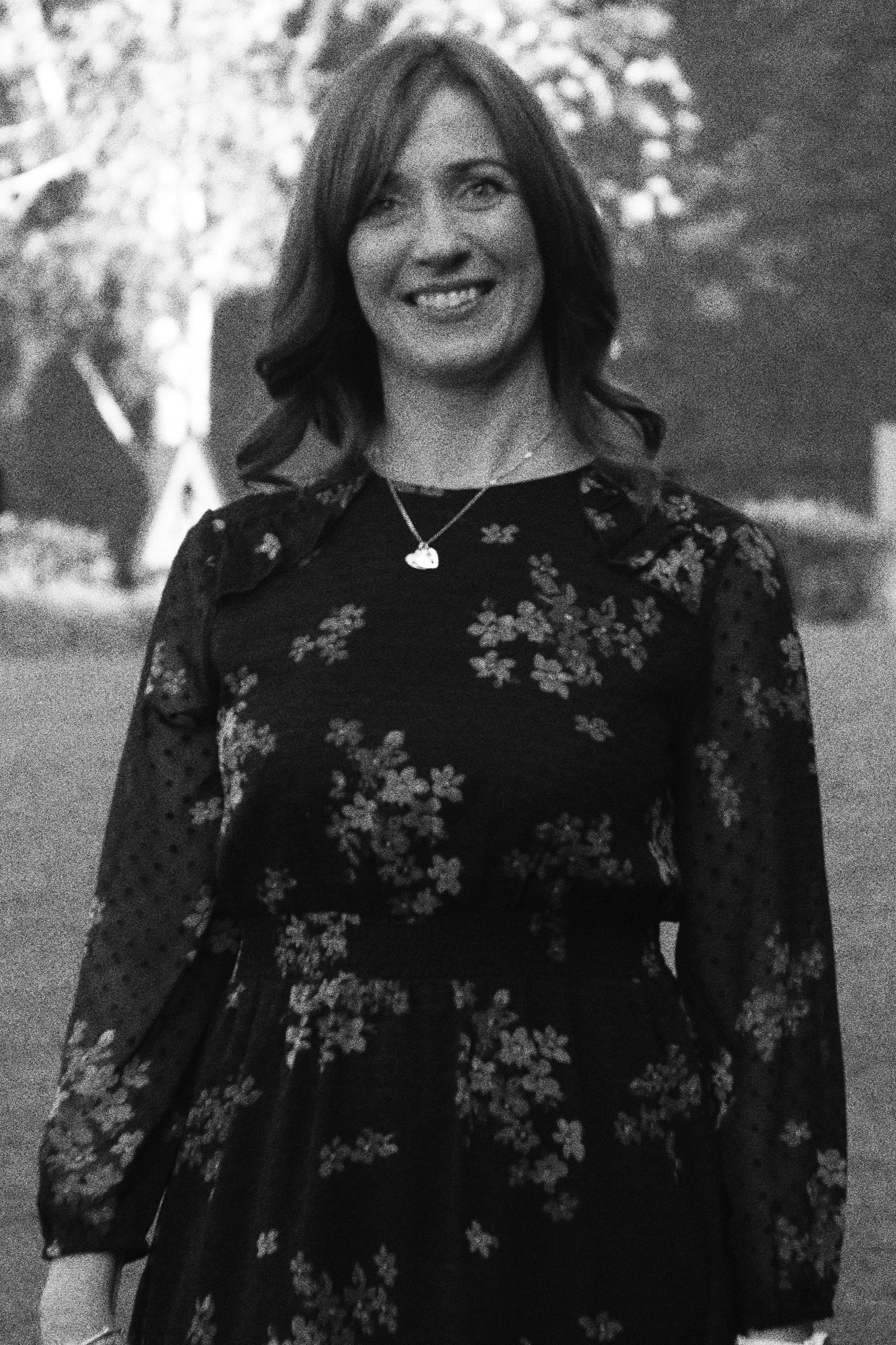 A woman with shoulder-length hair, smiling, wearing a floral dress and a necklace, standing outdoors.