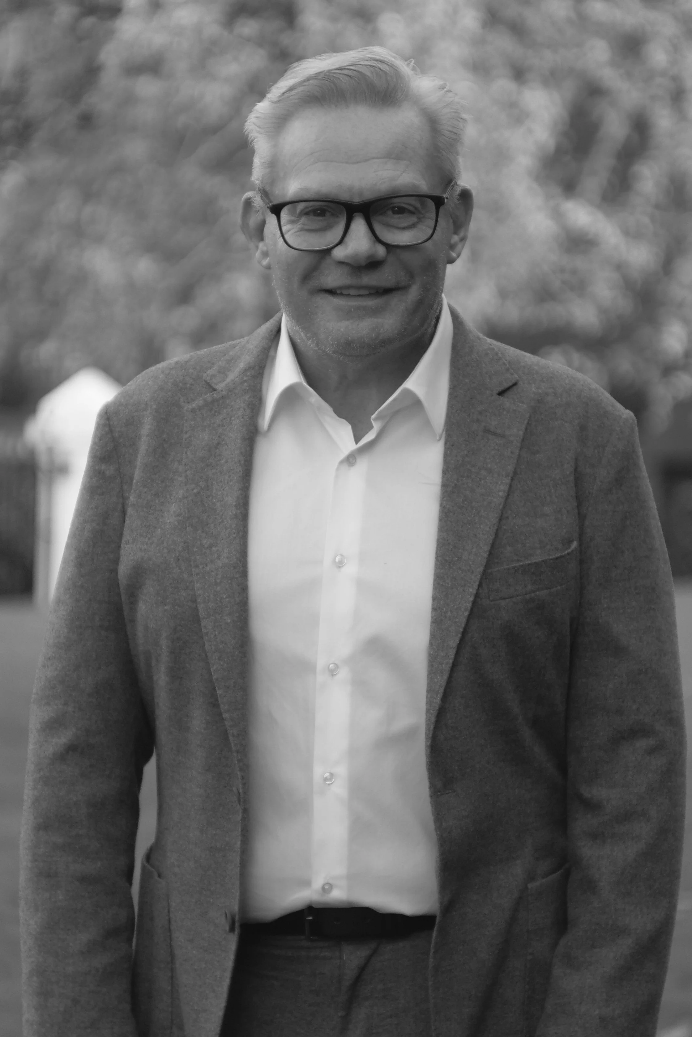 A middle-aged man with glasses, wearing a white shirt and a gray blazer, smiling outdoors.