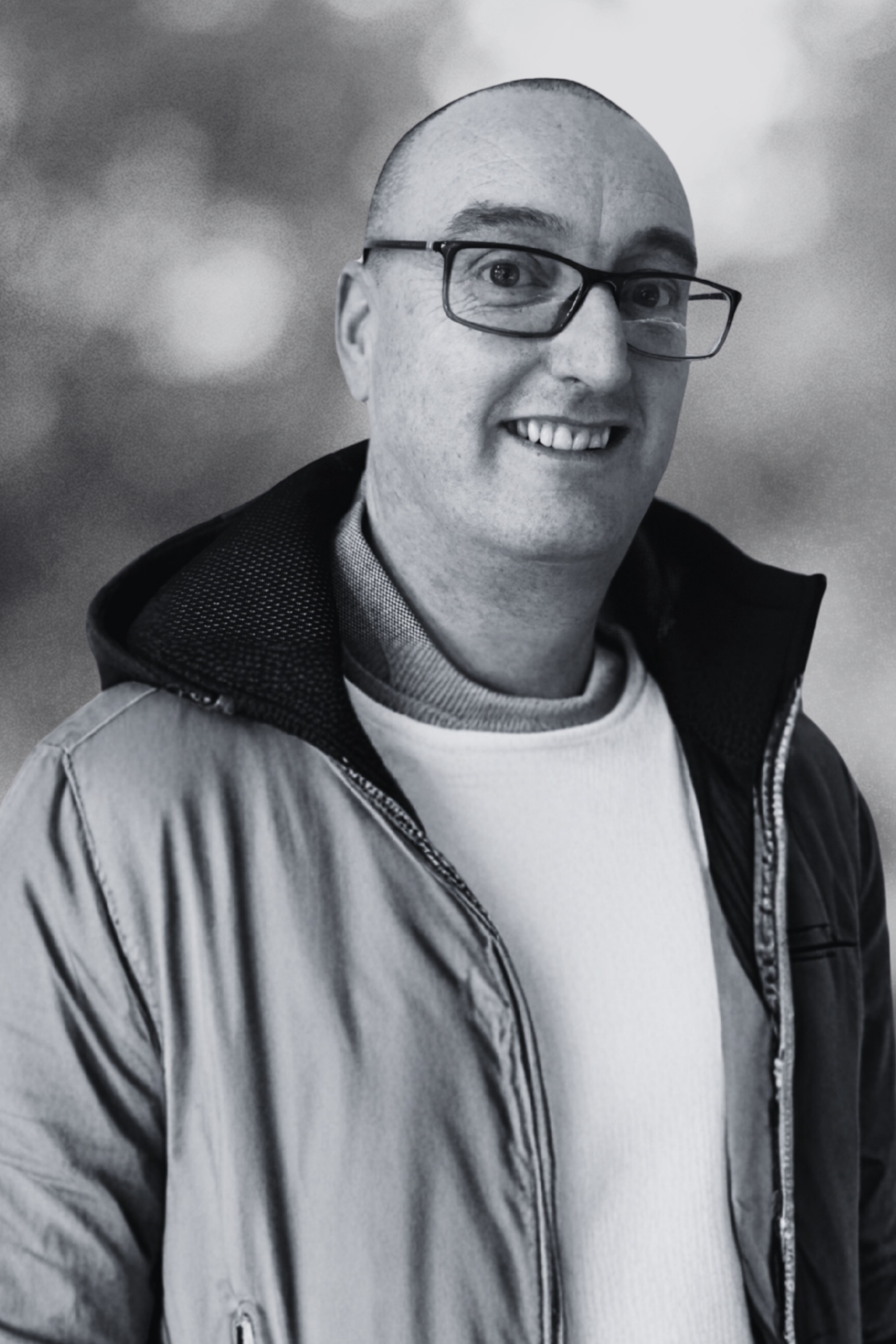 Black and white portrait of a smiling man with glasses, wearing a zip-up jacket over a sweater, against a cloudy background.