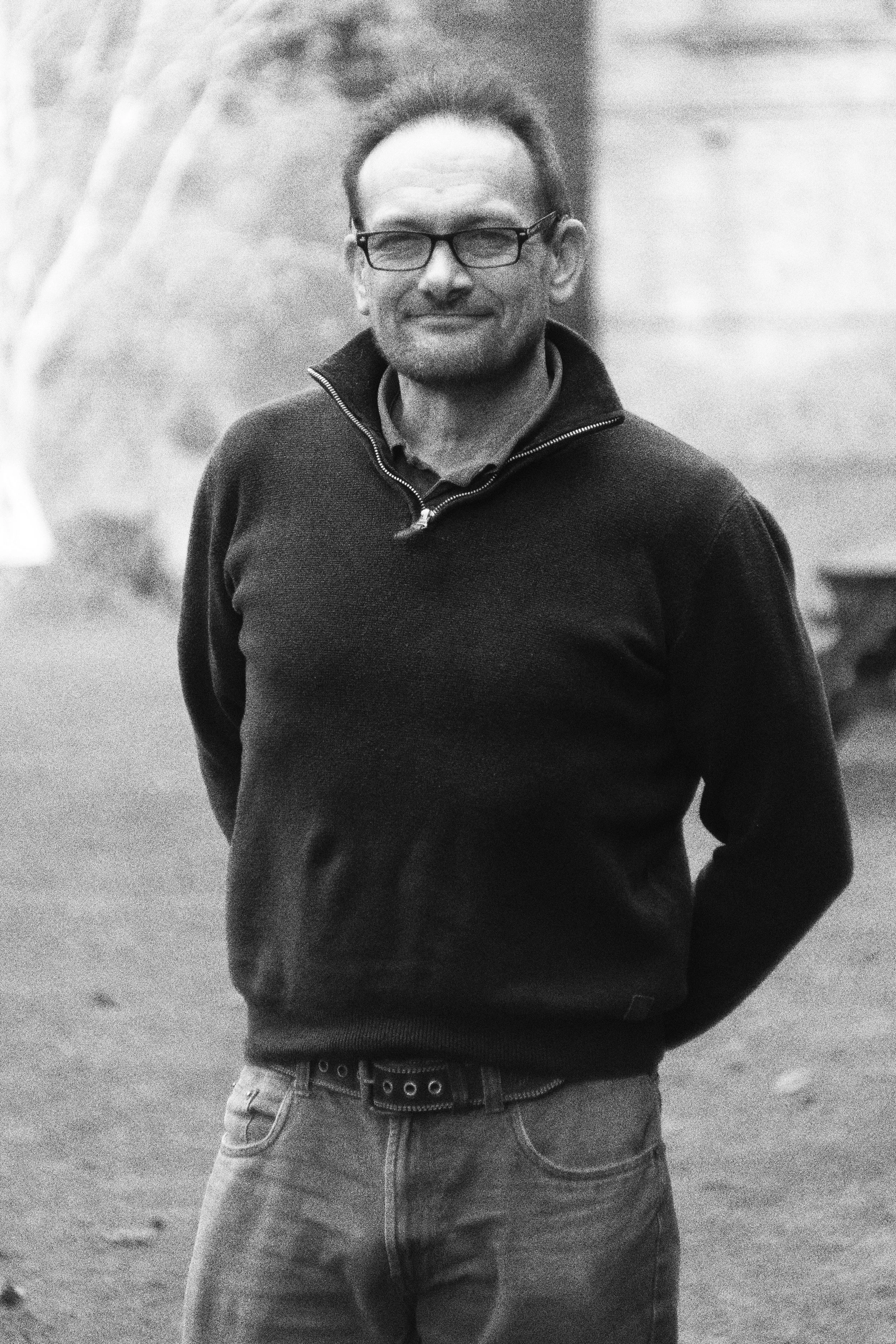 A man wearing glasses, a zip-up sweater, and jeans, standing outside with his hands behind his back, smiling slightly.