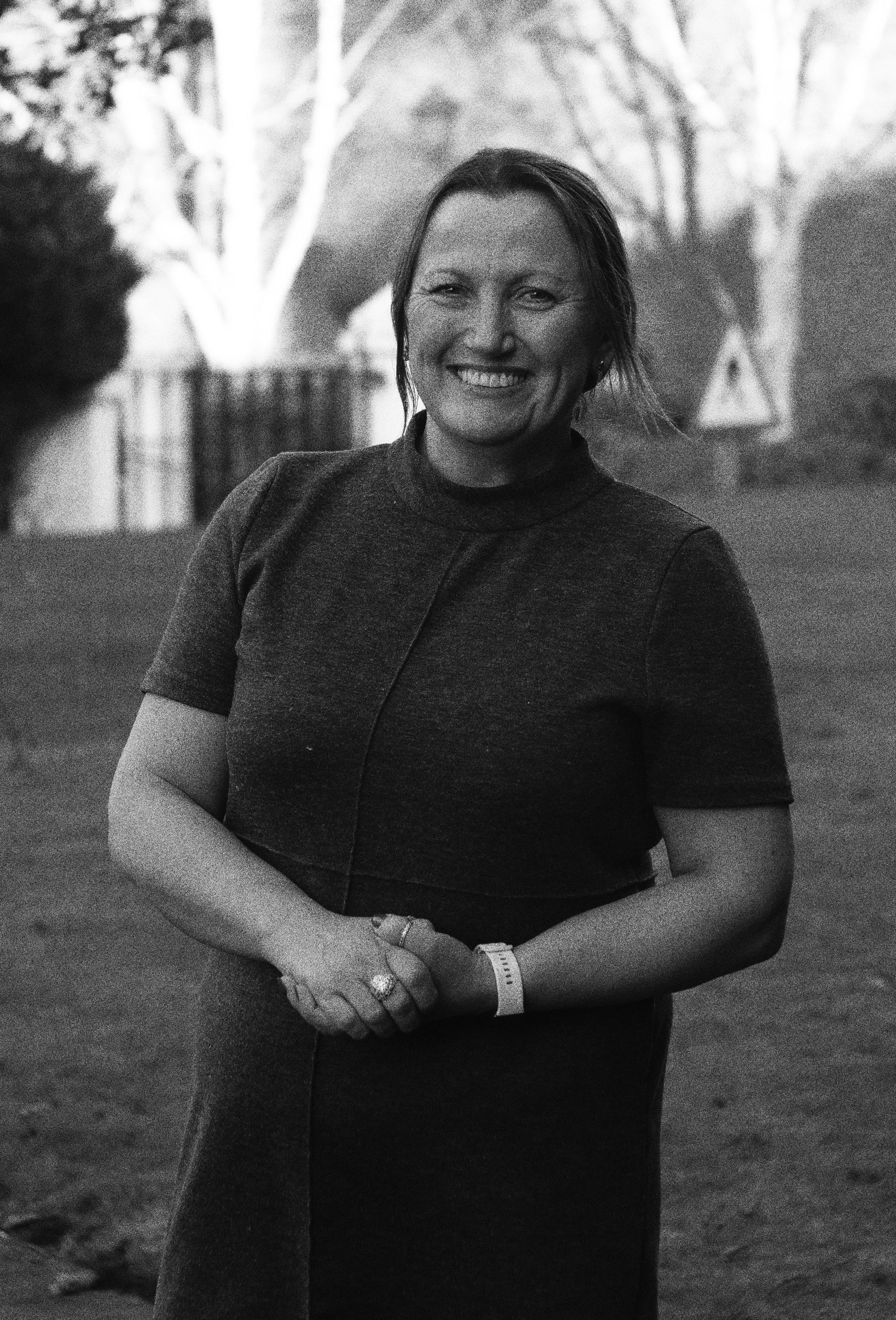A woman is smiling outdoors, wearing a dark short-sleeved top, with trees and a playground slide in the background.