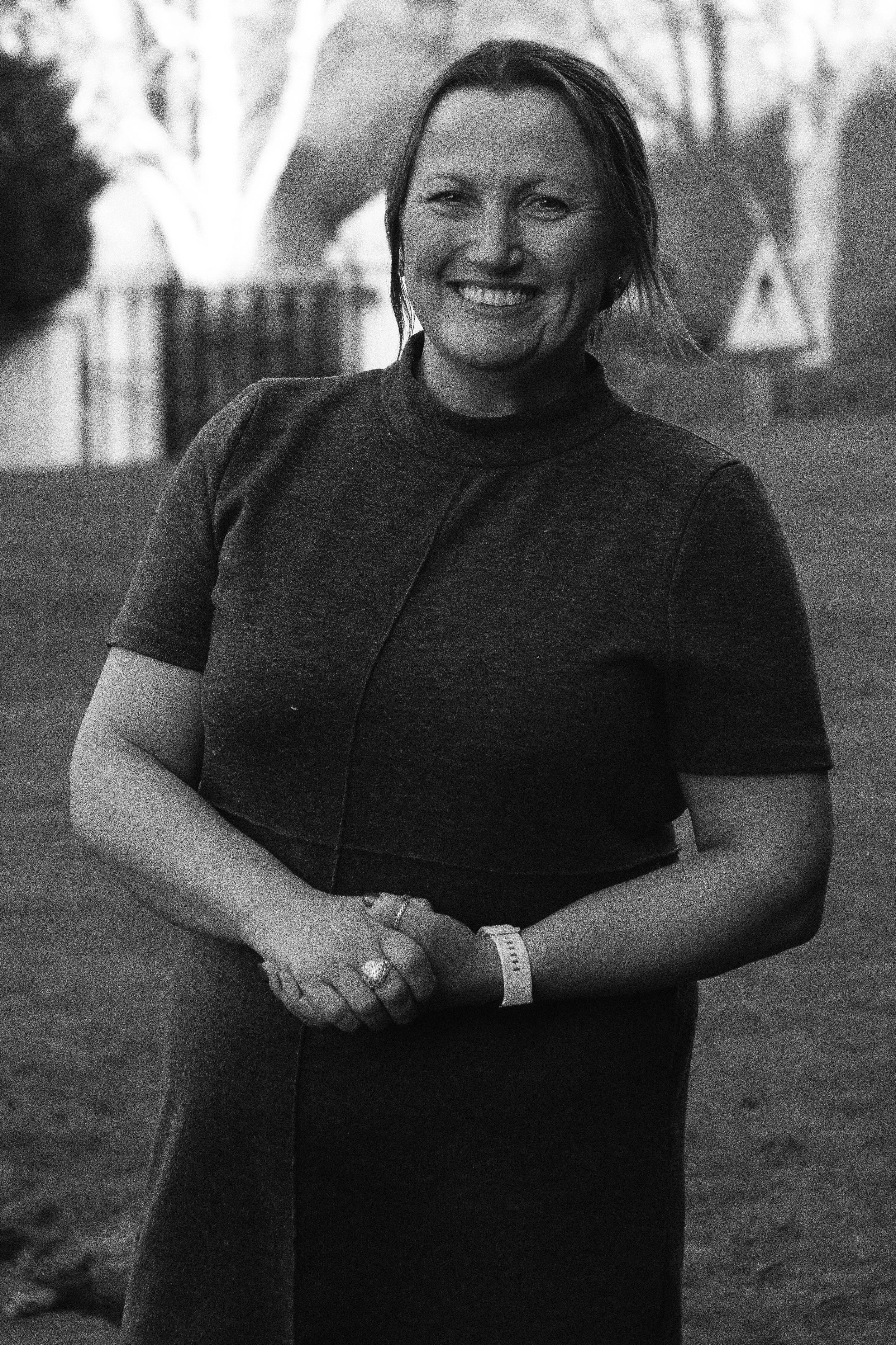 A smiling woman with short hair wearing a dark shirt, standing outdoors with trees and a fence in the background.