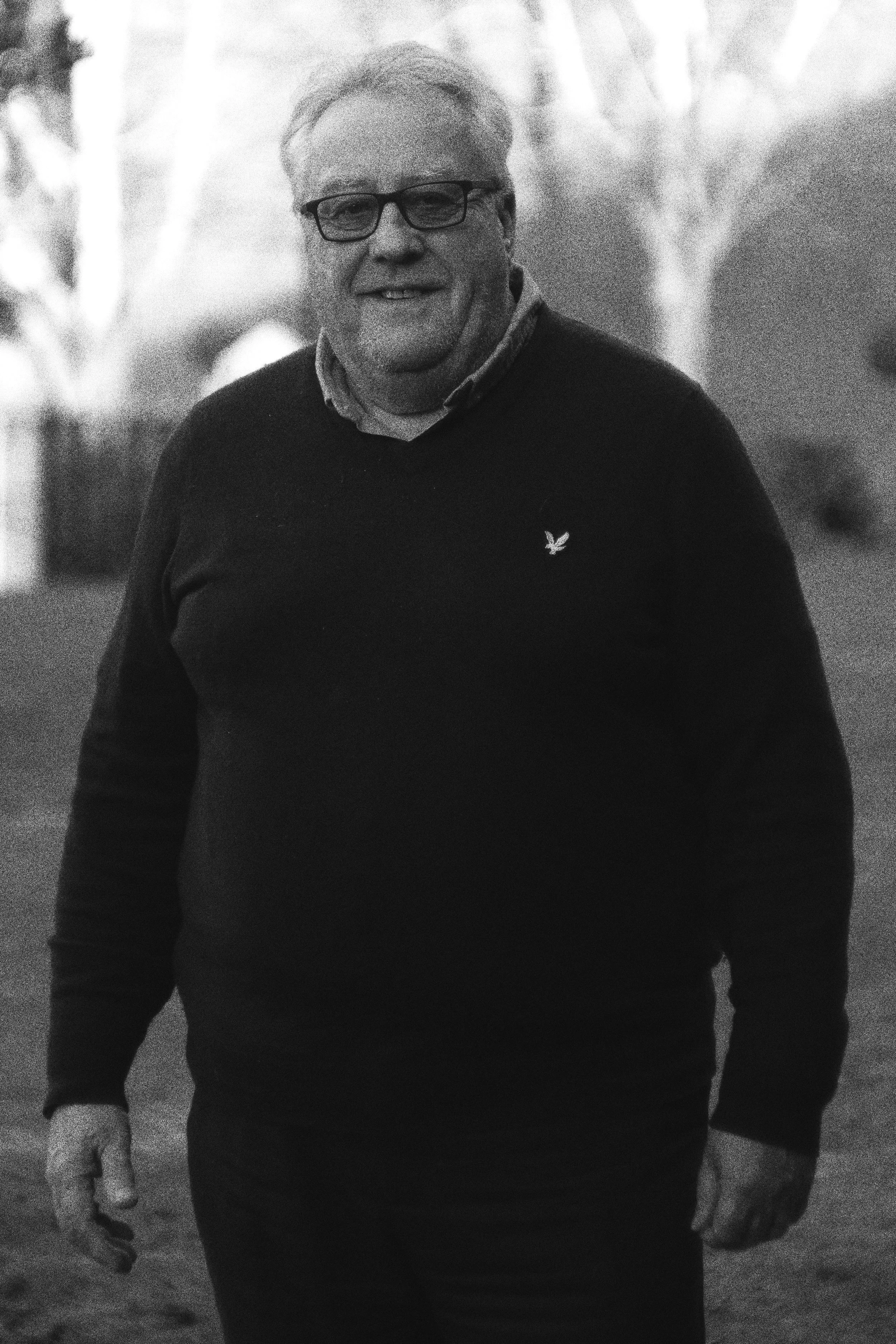 A smiling man outdoors in a casual outfit, wearing glasses and a dark sweater, with trees blurred in the background.
