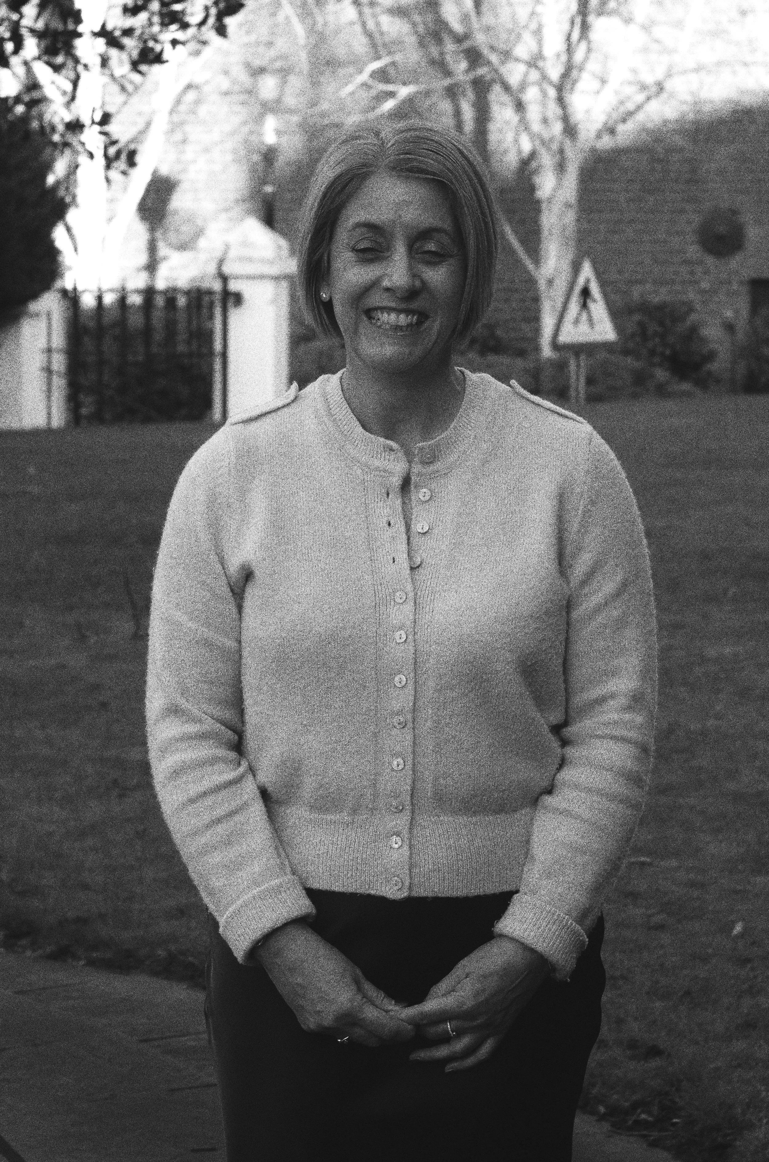 Black and white photo of a smiling woman with short hair, wearing a cardigan and placing her hands in front of her, outdoors in a park or garden setting.