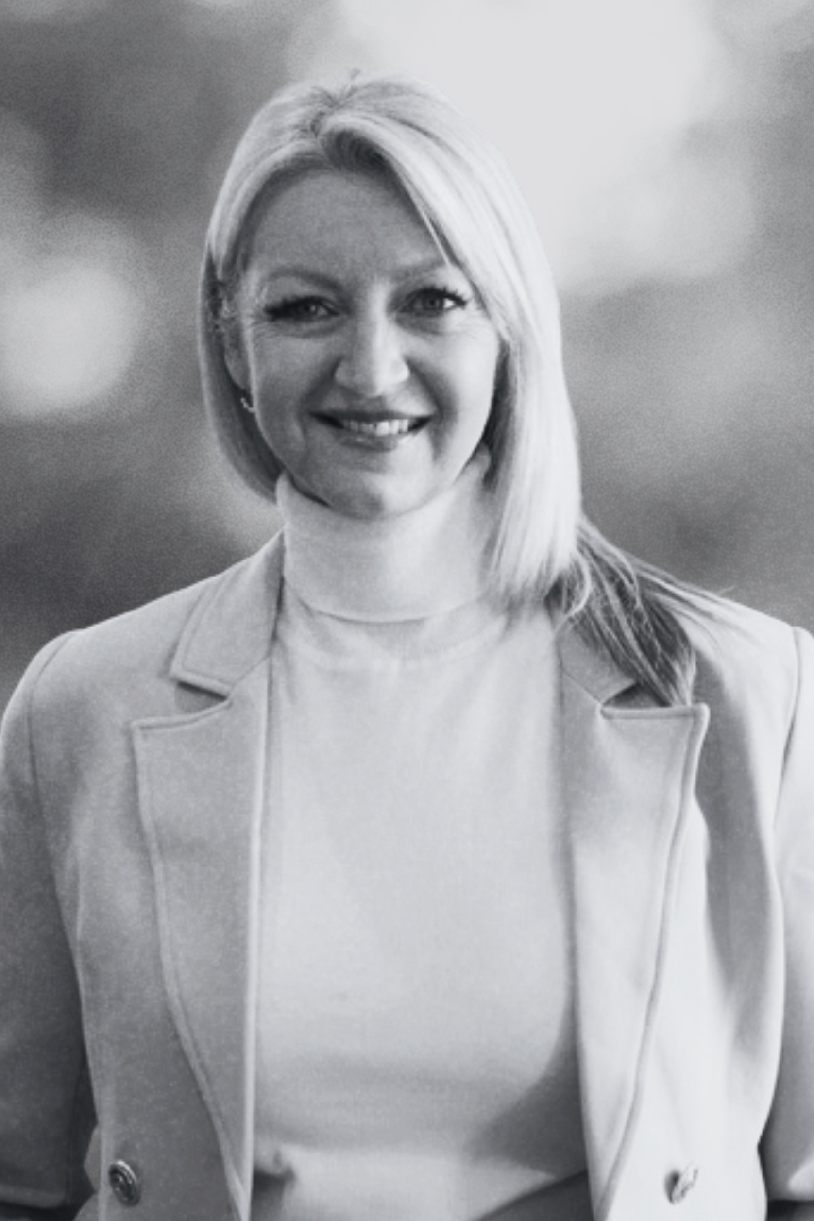 Black and white portrait of a smiling woman with shoulder-length hair, wearing a light blazer and turtleneck sweater, with a blurred outdoor background.