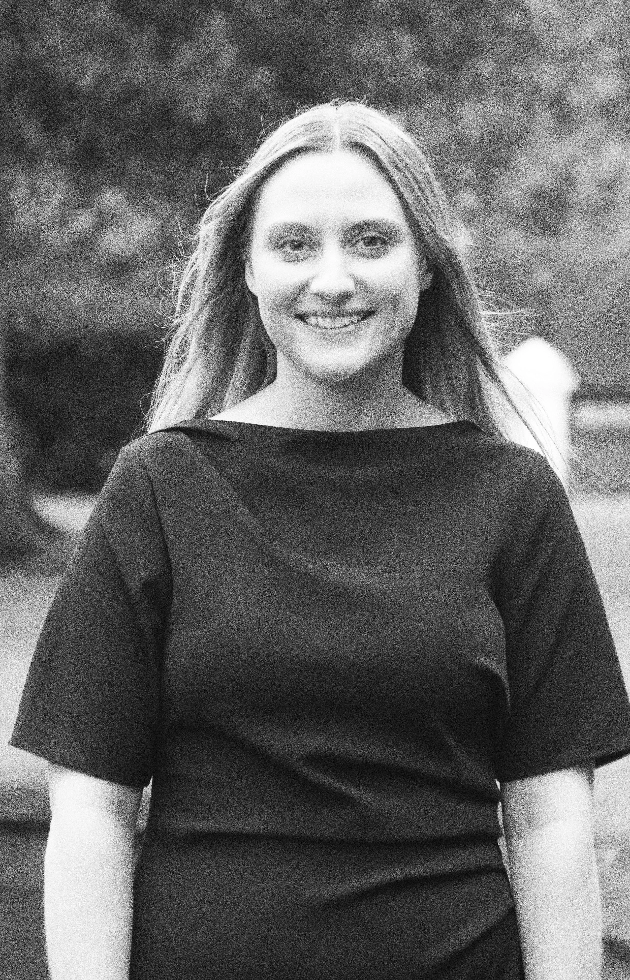 Black and white portrait of a young woman outdoors, smiling, with long hair, wearing a short-sleeved dark top.