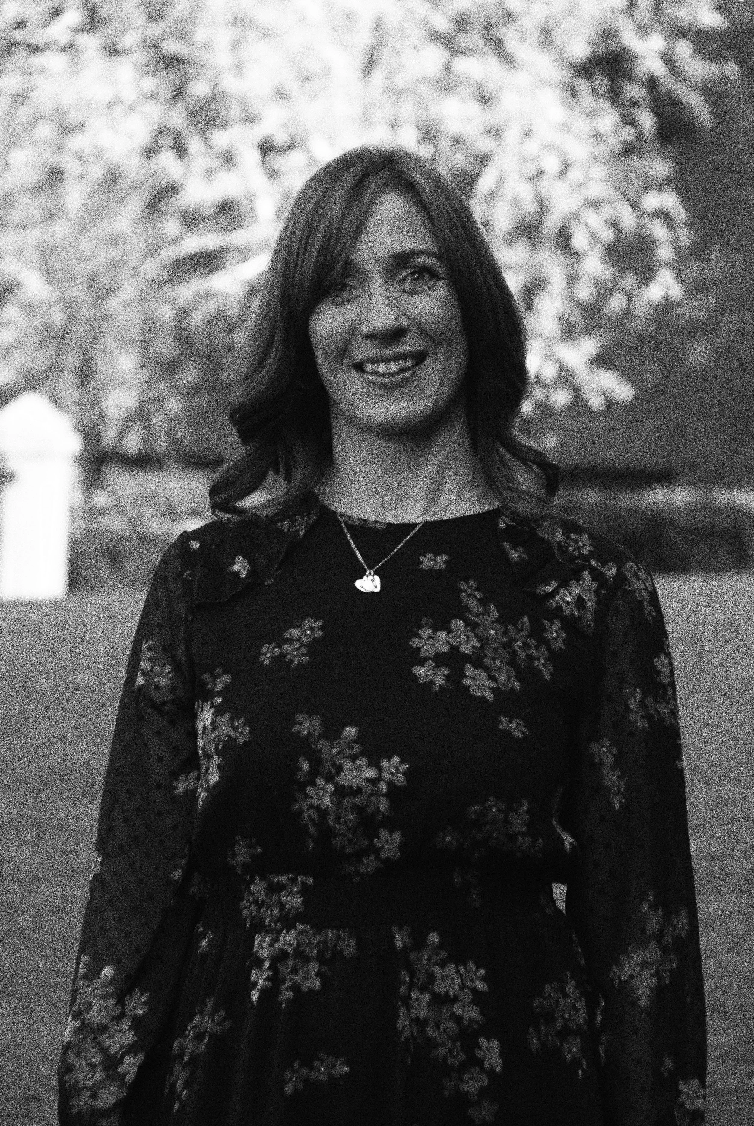 A woman smiling outdoors, wearing a floral dress and a necklace with a heart-shaped pendant.