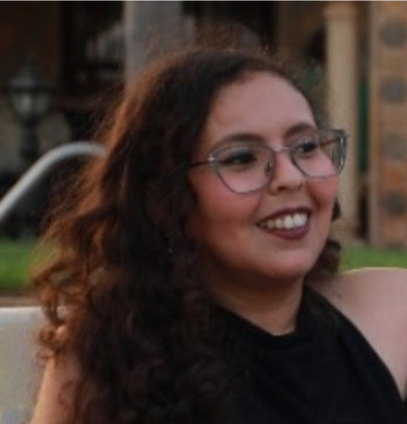 Jeune femme souriante avec lunettes, cheveux bouclés, de couleur brune, portant un haut noir, assise en extérieur