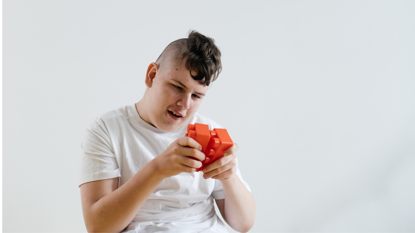 Un garçon autiste regarde un puzzle rouge avec confusion, assis contre un mur blanc.