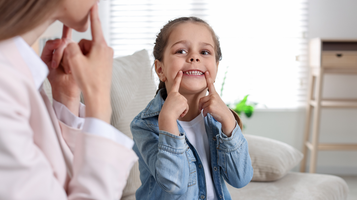 Une petite fille souriante lors d'un rendez-vous avec son orthophoniste, tirant la commissure de ses lèvres pour articuler le i.