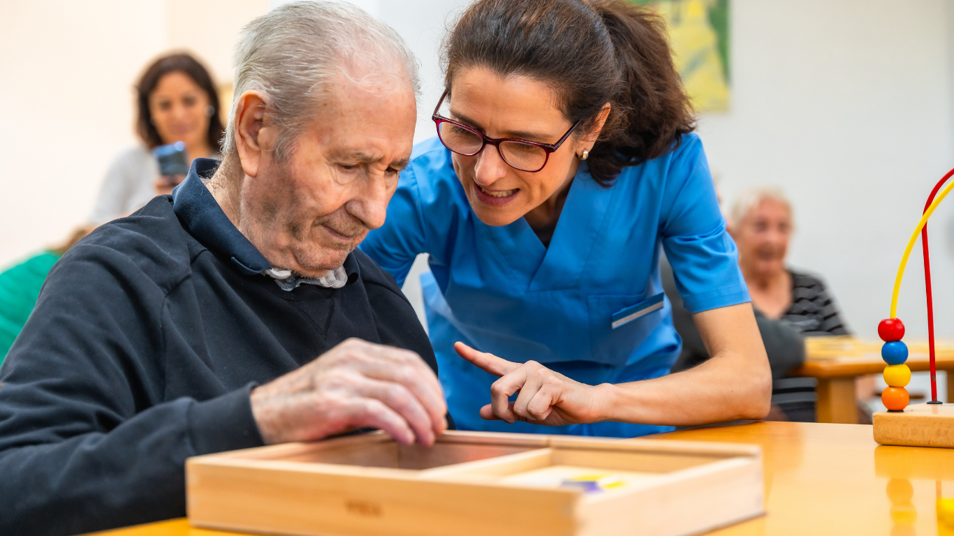 Une orthophoniste aide un homme âgé à jouer à un jeu de société dans une salle de soins, avec d'autres personnes en arrière-plan