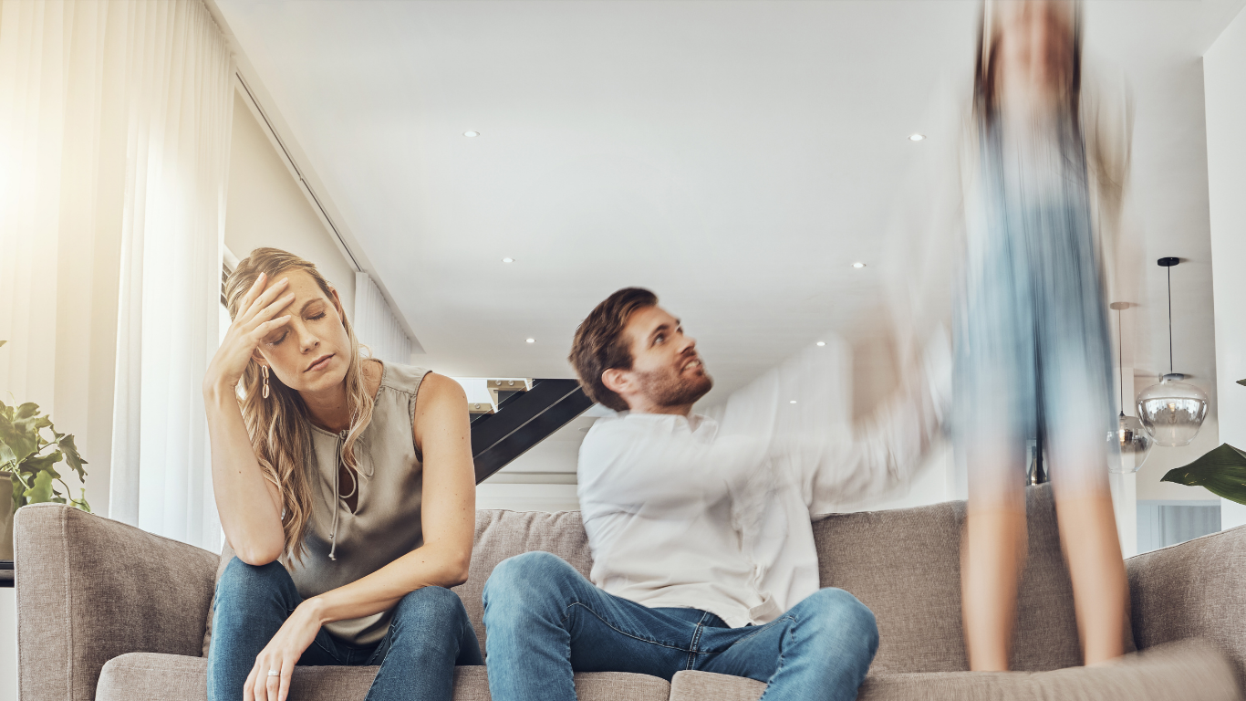 Des parents assis sur un canapé dans un salon lumineux, le papa regarde son enfant qui saute.