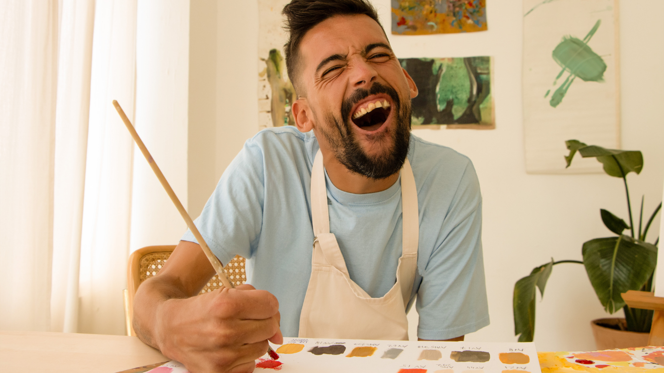 Un homme souriant et riant, portant un tablier blanc, tient un pinceau et semble peindre ou s'amuser dans un studio d'art.