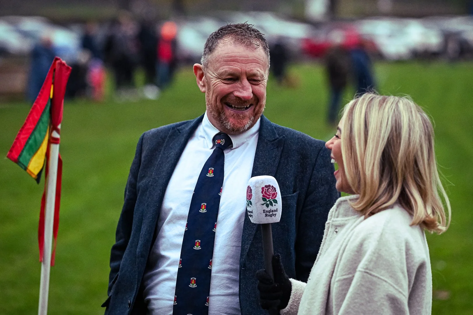 A man in a suit and tie being interviewed outdoors with a woman holding a microphone marked 'England Rugby.' Both are smiling and laughing, with a grassy field and parked cars in the background.
