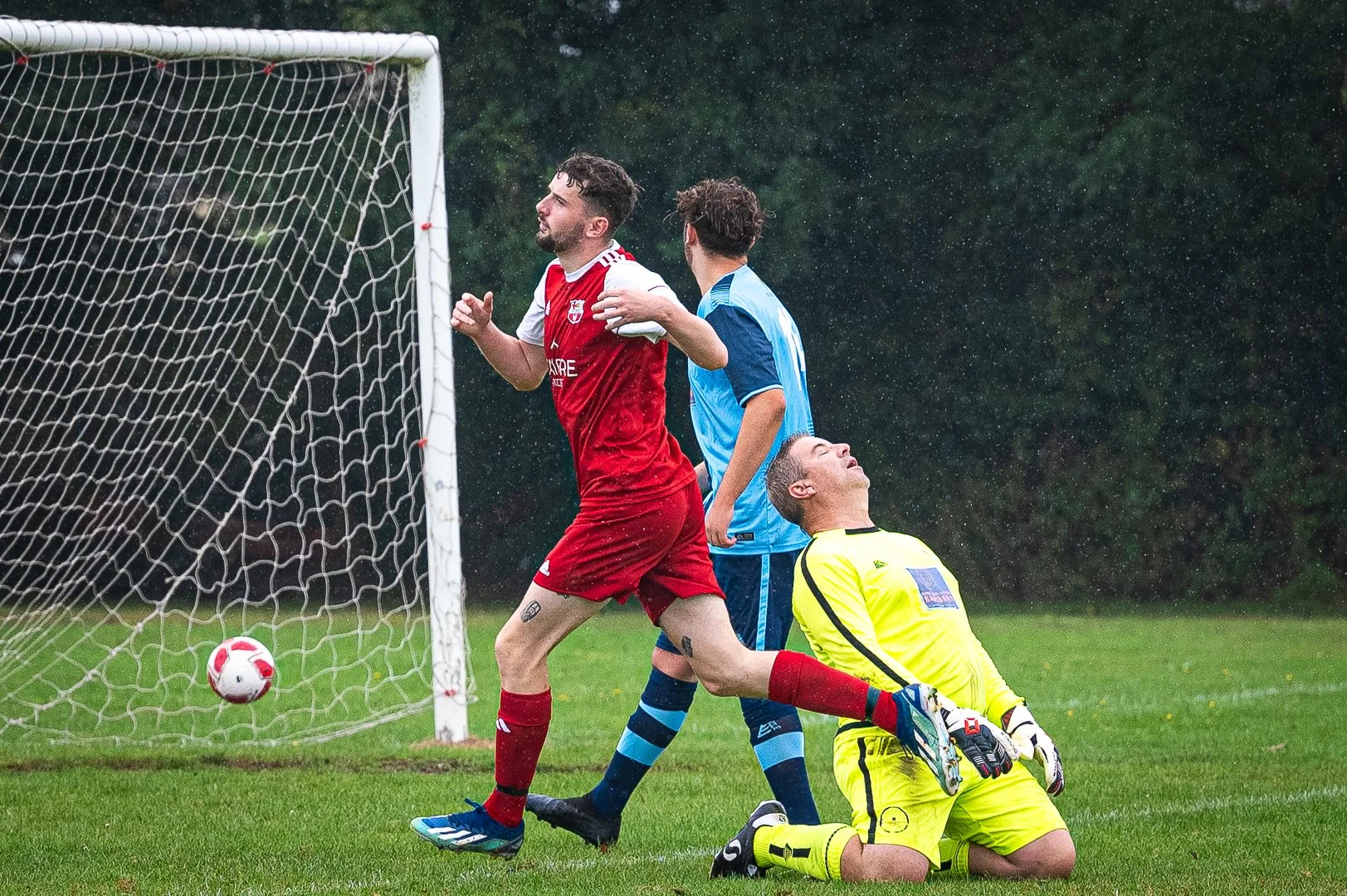 A soccer game with players near the goal; one player in red is celebrating, another in blue is standing, and the goalkeeper in yellow is on his knees after a missed scoring attempt.