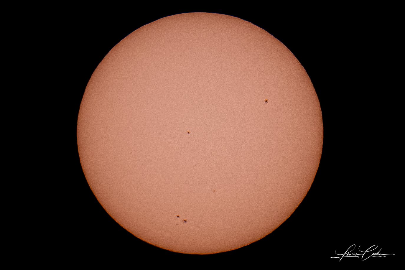 Close-up photograph of the sun with several dark sunspots visible against its orange surface.