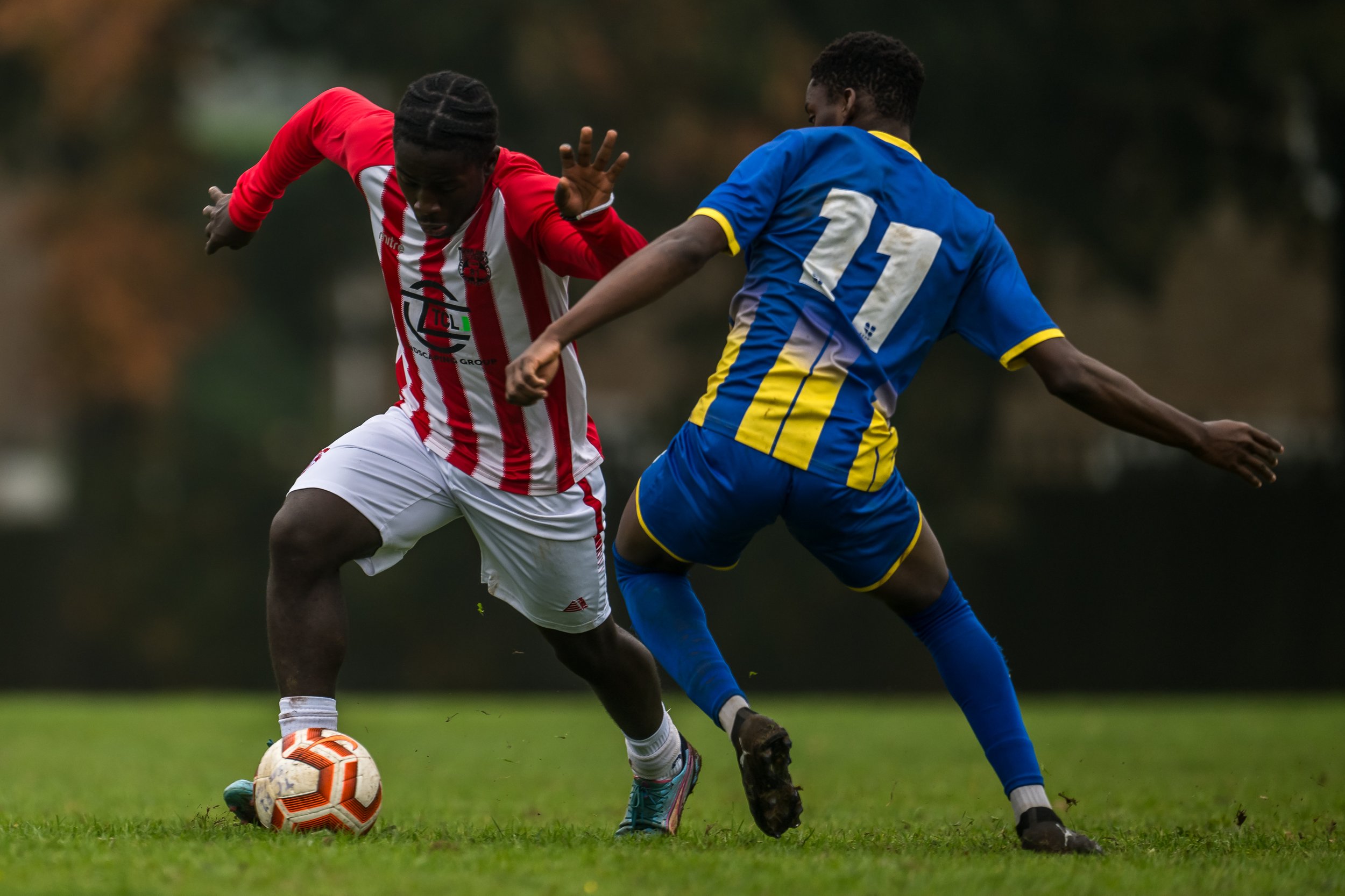 Two soccer players competing for ball on field, one in red and white striped jersey, the other in blue and yellow jersey.