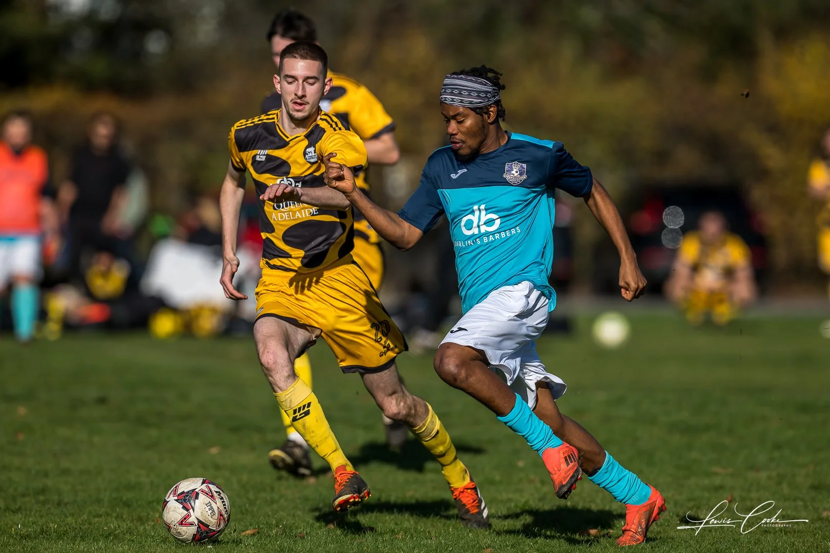 Two soccer players compete for the ball on the field during a match, with other players and spectators in the background.