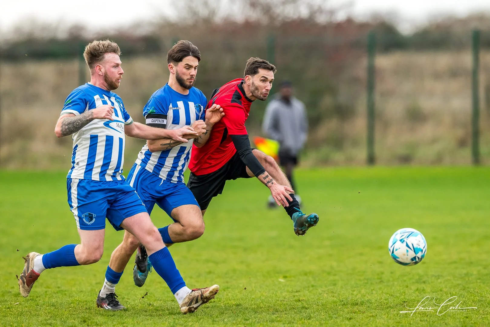 Three men playing soccer on a grass field, two in blue and white uniforms and one in red and black, chasing after a soccer ball during daytime.