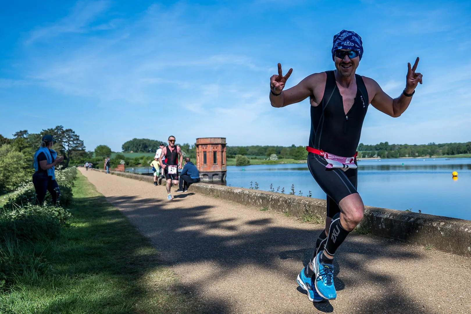 A man wearing a black athletic suit, blue running shoes, a blue head wrap, and sunglasses is running along a scenic lakeside path, making peace signs with both hands and smiling. In the background, other runners and spectators are near a red brick st