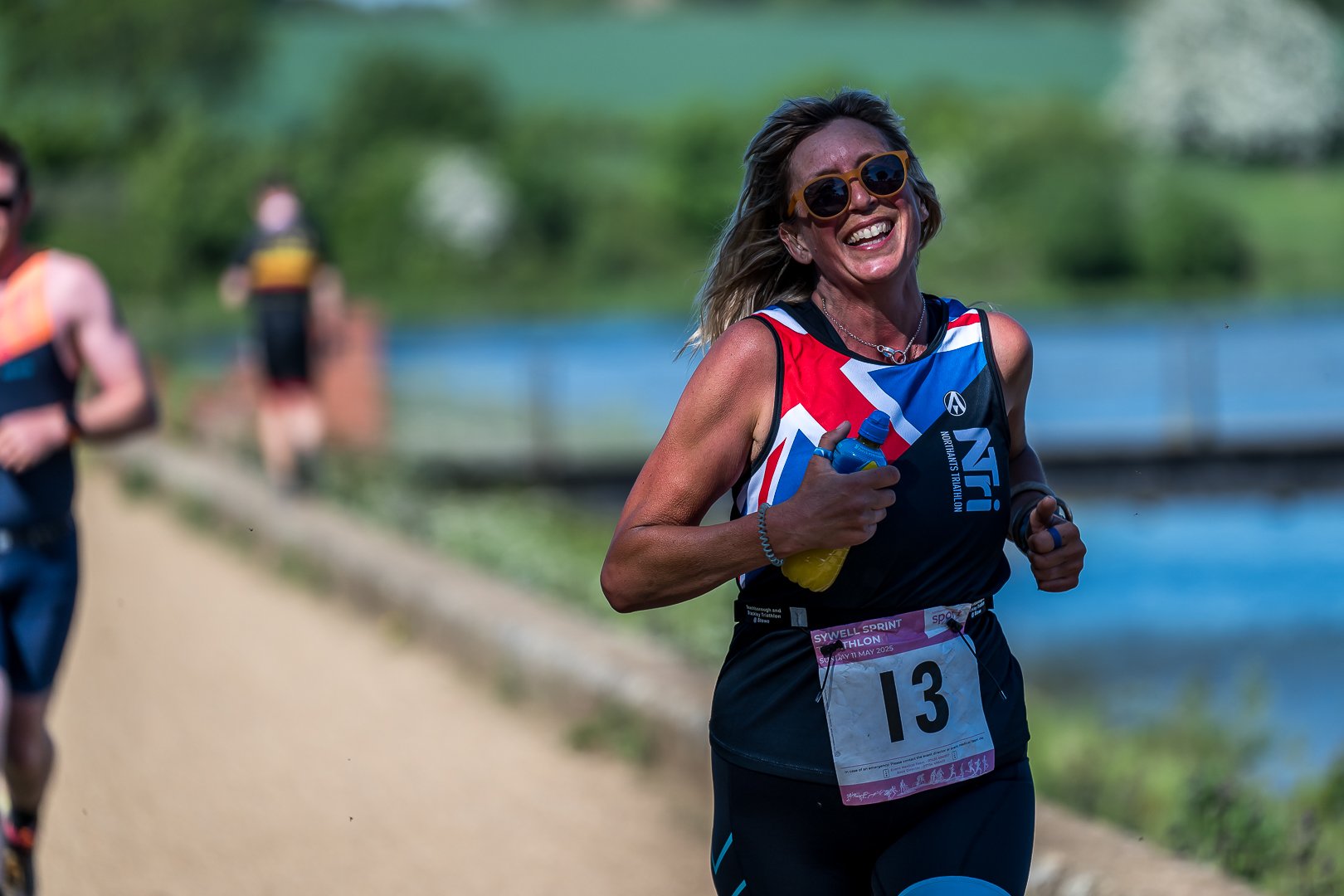 Smiling female runner with sunglasses holding water bottle during outdoor race, wearing a race bib numbered 13.