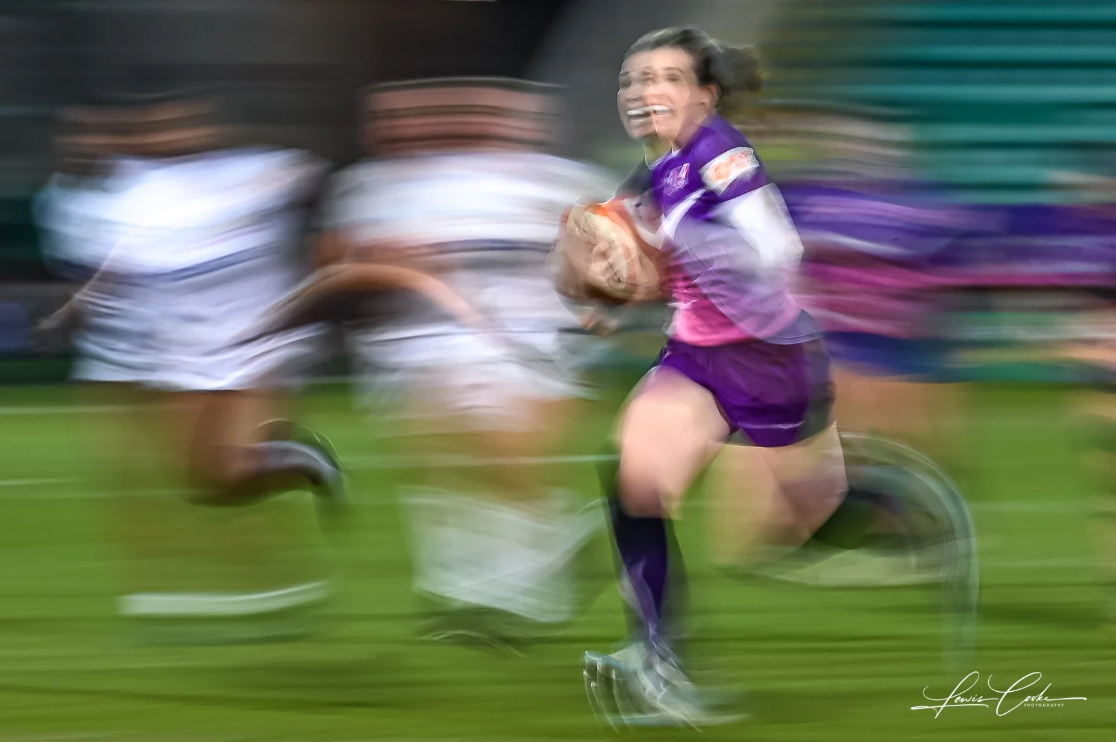 A female rugby player running with a rugby ball during a match, with blurred background showing other players on a grass field.