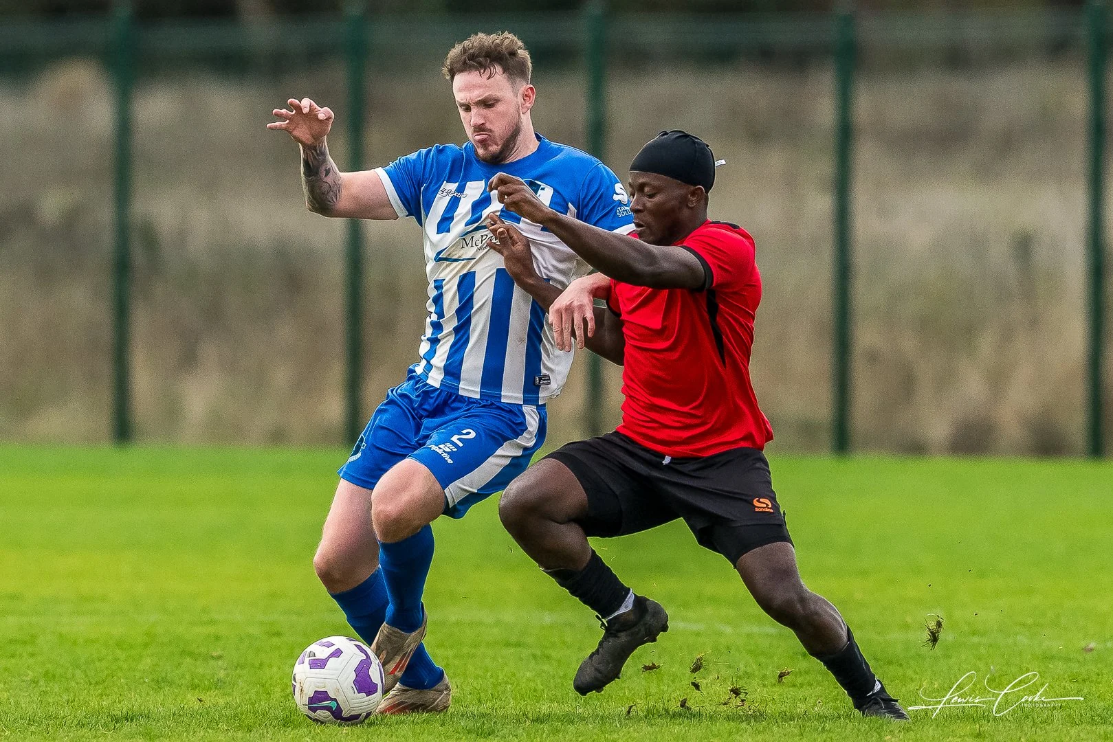 Two soccer players competing for the ball on a grassy field, one in a blue and white striped uniform, the other in a red shirt and black shorts, in an outdoor setting.