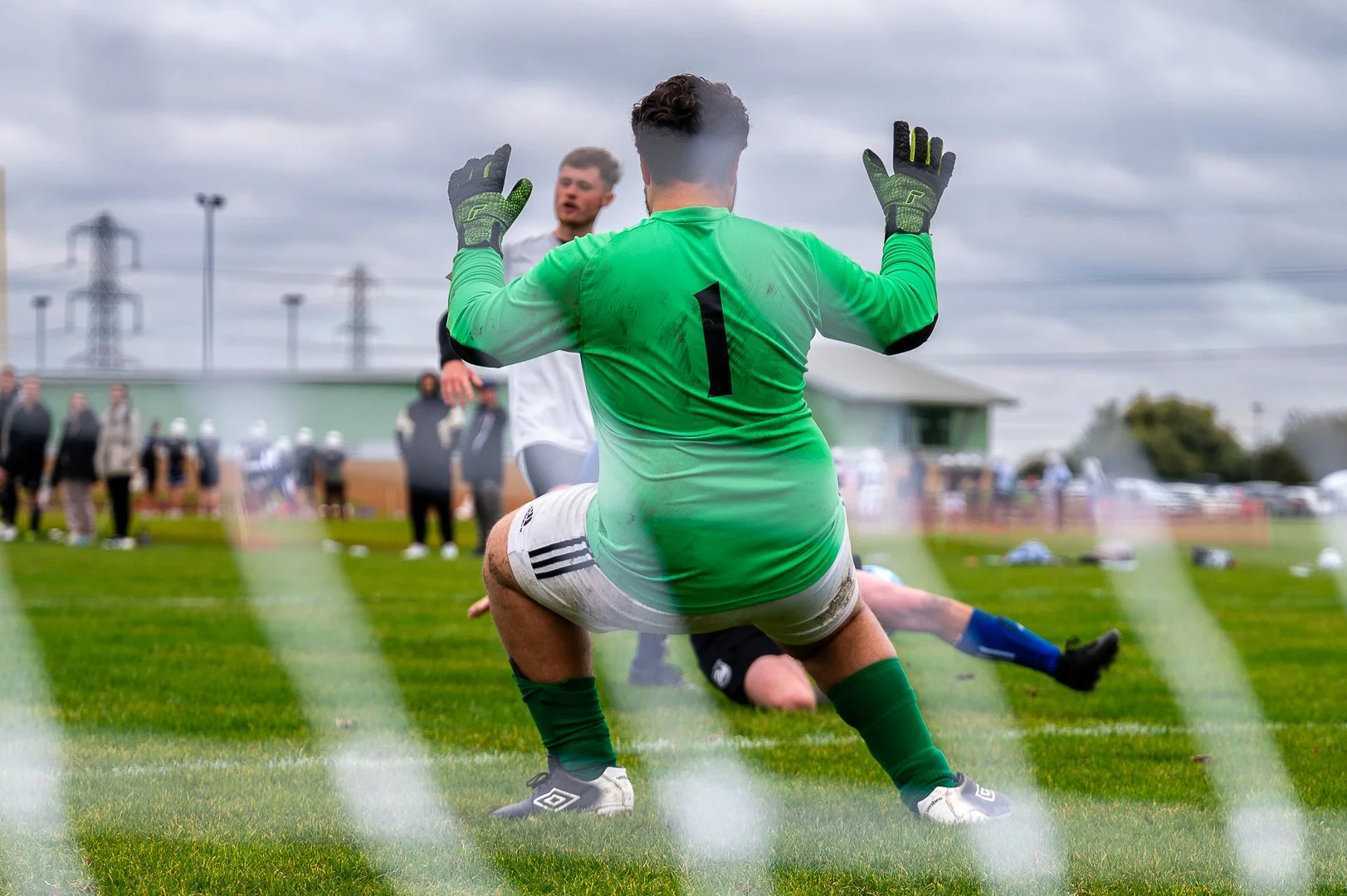 Soccer goalkeeper in green jersey with number 1, gloves, and shorts kneeling on the grass with arms raised during a game, with players and spectators in the background under cloudy skies.