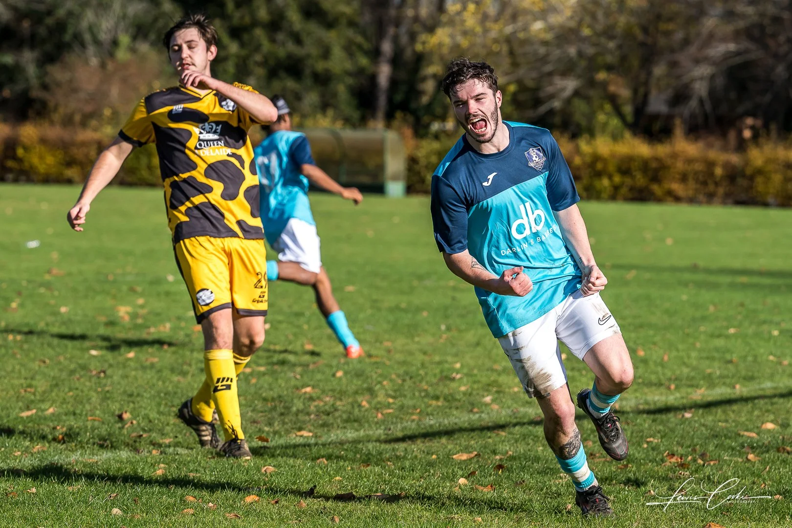 A soccer player in a blue shirt and white shorts celebrating on a grassy field. Two other players in blue and yellow uniforms are in the background. Autumn trees are visible in the distance.