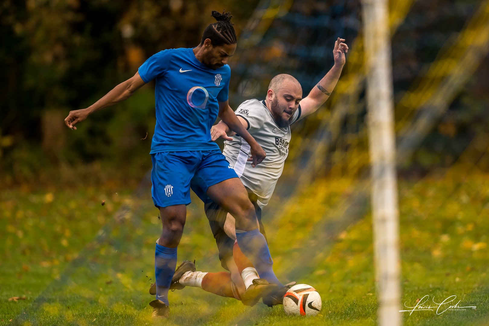 Two soccer players competing for the ball during a match, outdoors on a grass field with trees in the background.