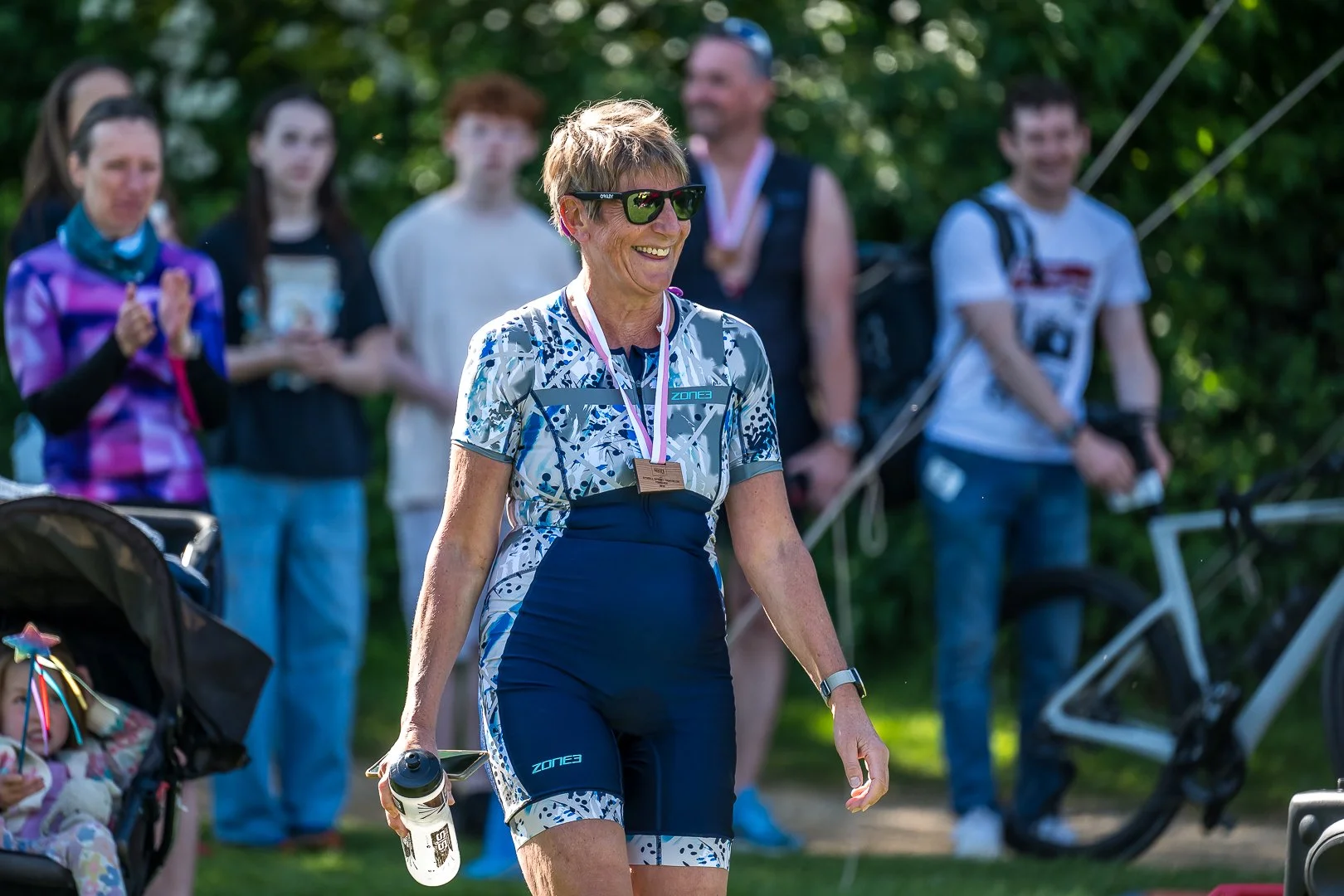 A smiling woman in cycling gear walking with a water bottle, surrounded by a group of people outdoors.