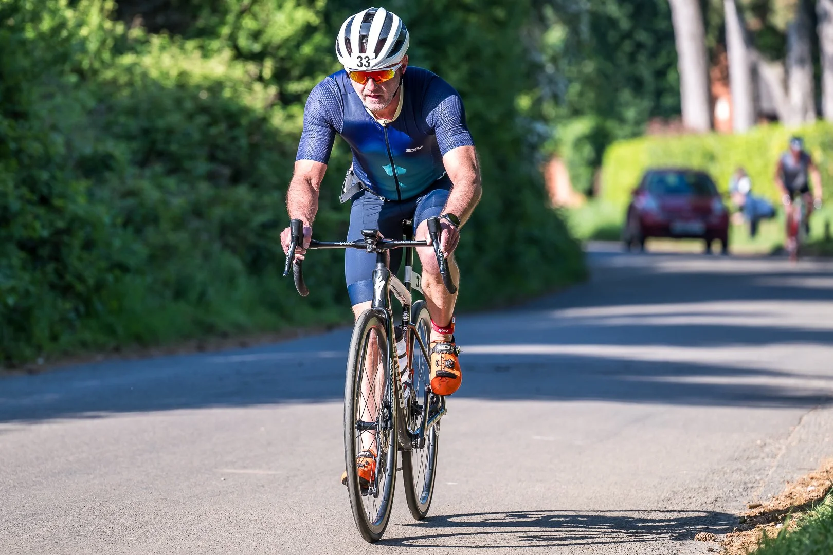 A man riding a bicycle on a paved road surrounded by green trees, wearing a blue cycling kit, helmet, sunglasses, and orange shoes.