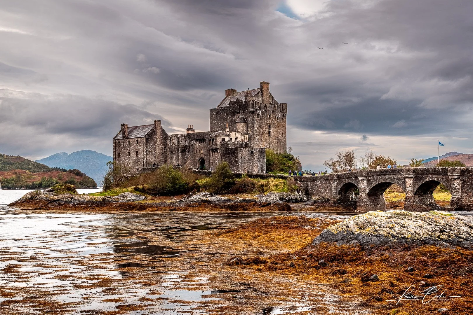 A medieval stone castle with a moat and arched stone bridge under a cloudy sky, surrounded by a landscape of mountains and water.