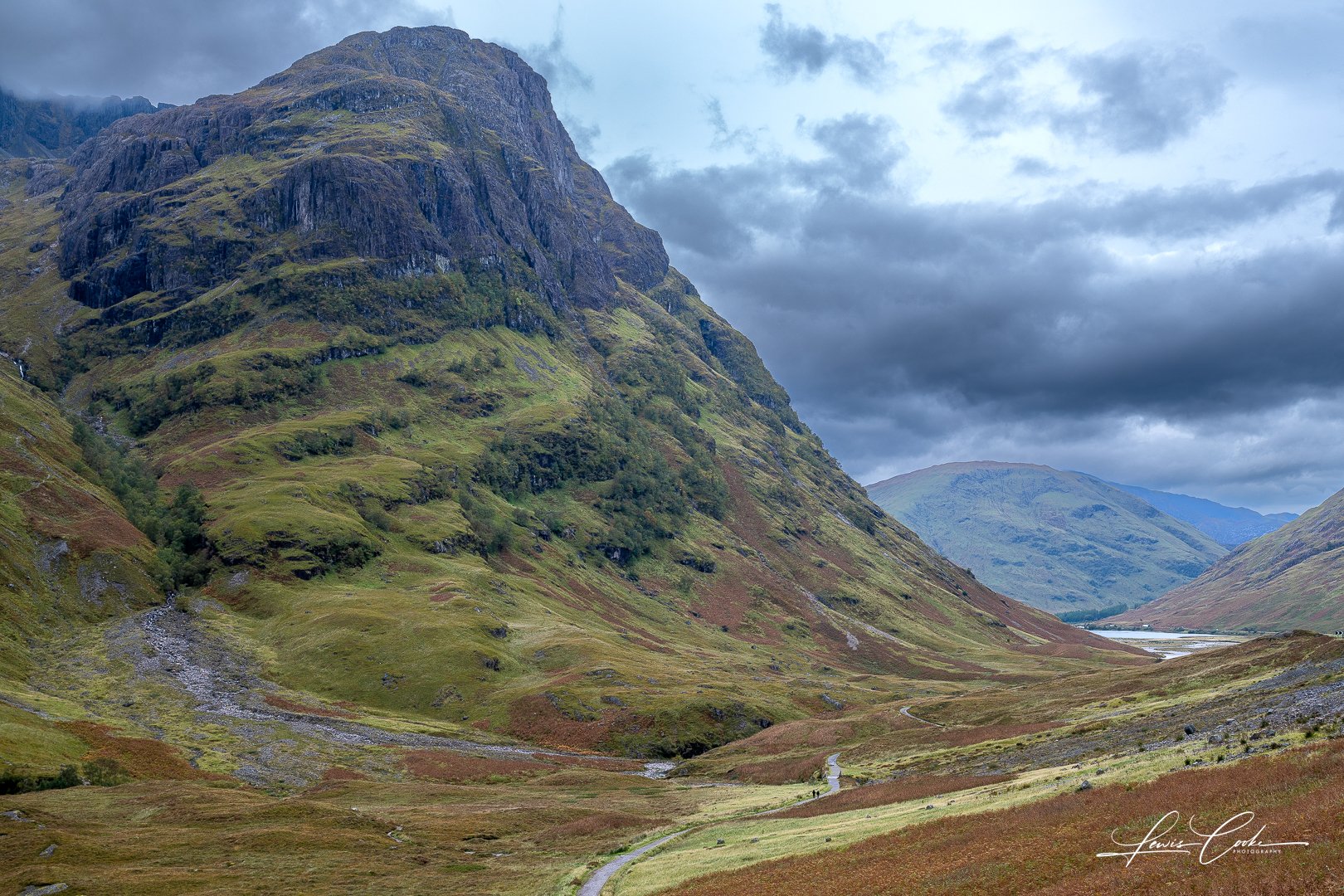 Scenic view of green mountains and a winding dirt trail in a valley under cloudy sky.