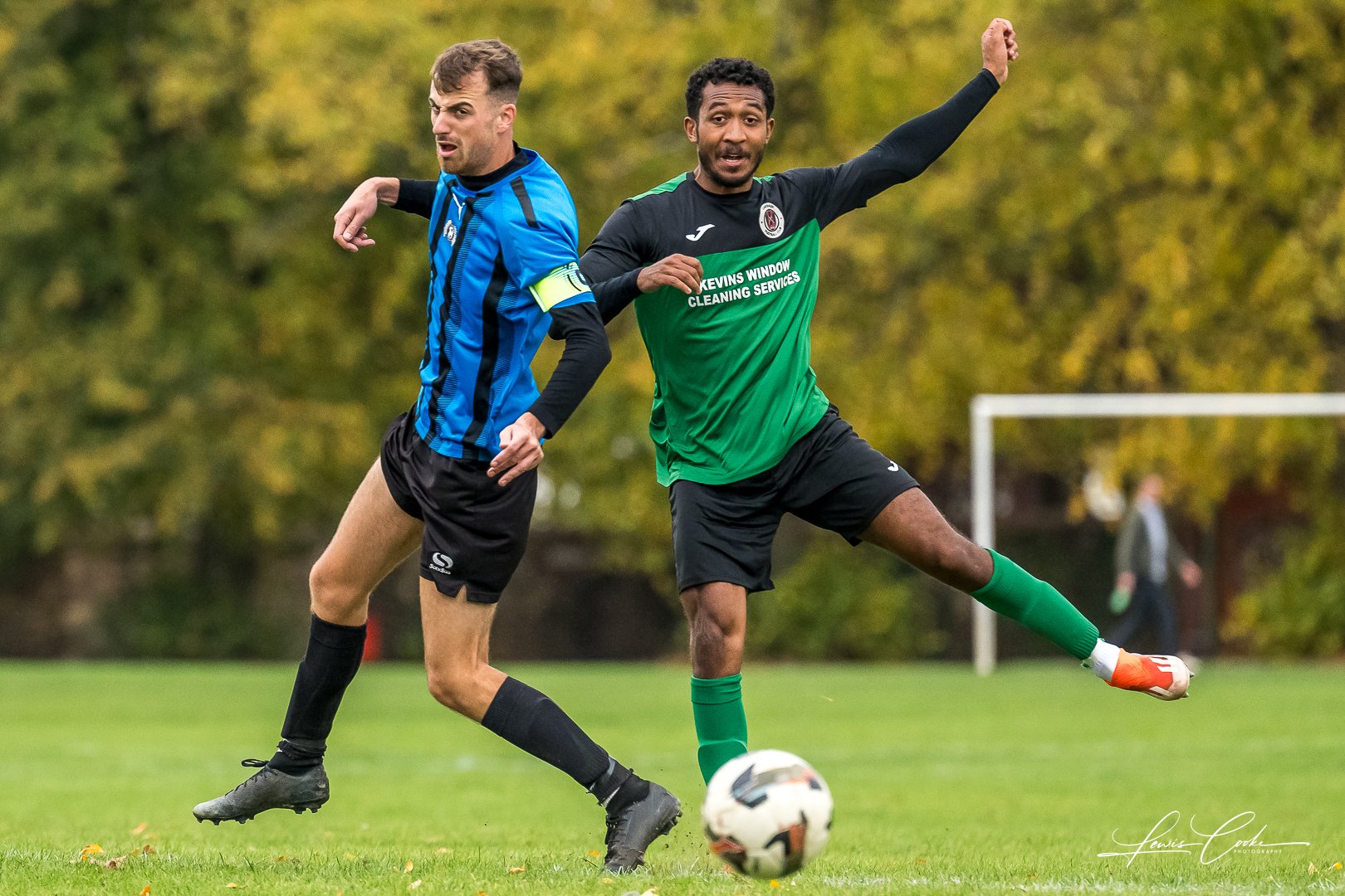 Two soccer players competing for the ball on a grassy field, with trees in the background.