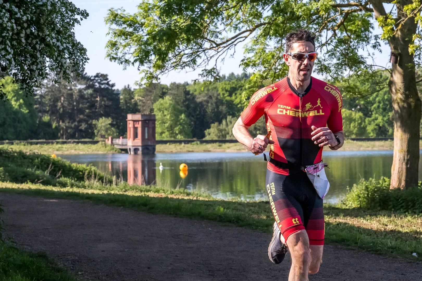 A man running in a park beside a lake, wearing sunglasses and a red and black athletic outfit with the words "Team Cherwell" on it.
