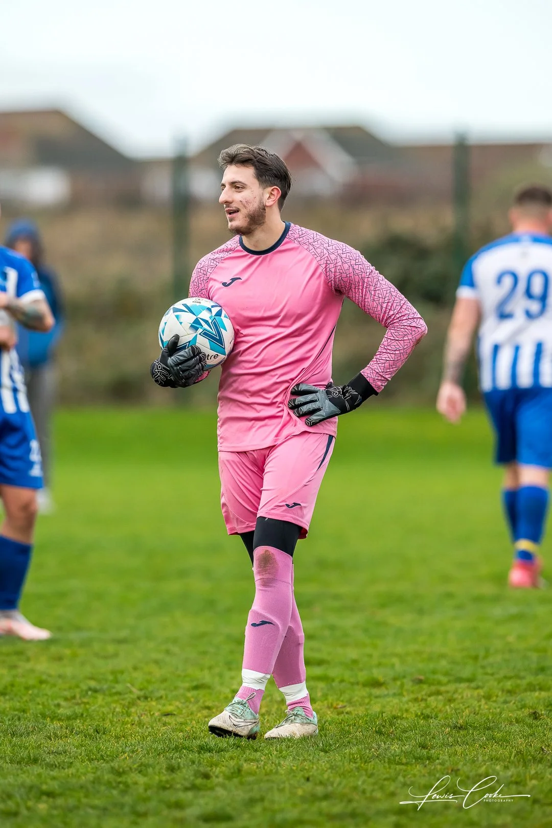 A male soccer goalkeeper in a pink uniform holding a soccer ball on a grassy field during a match, with other players in blue and white uniforms visible in the background.