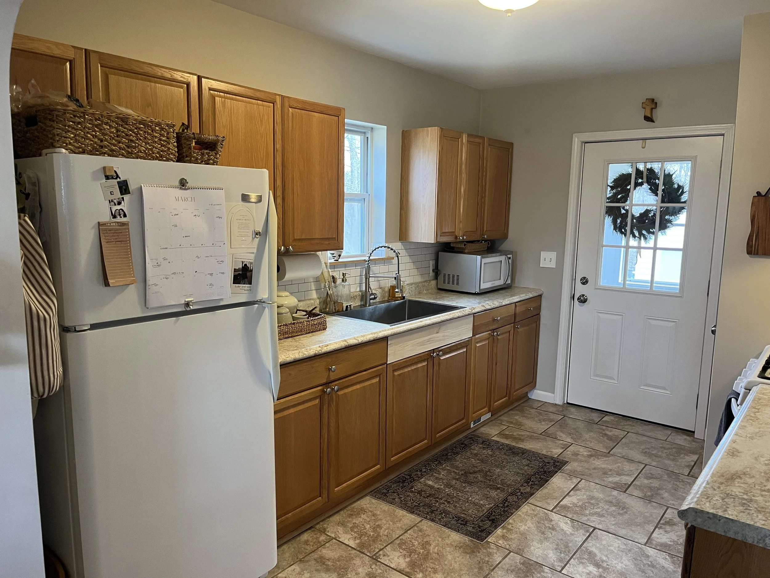 Kitchen with wooden cabinets, a white refrigerator with a calendar and photos, a window, a microwave, a paper towel holder, and a door with a wreath and a small cross above it.