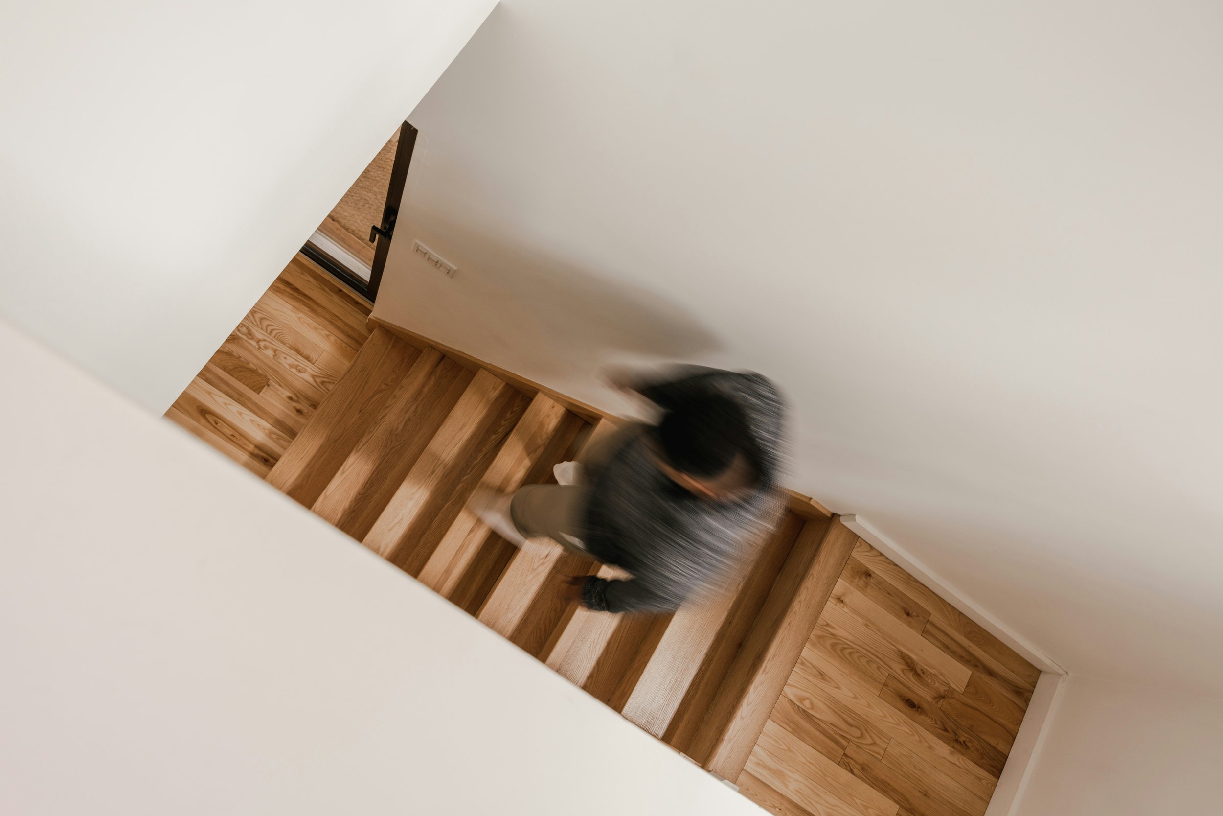 Blurred top-down view of a person walking down wooden stairs, with white walls and a door at the bottom.