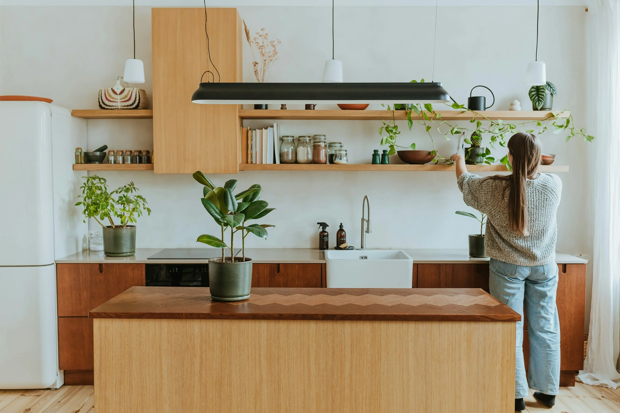 A woman with long hair wearing a beige sweater and blue jeans arranging plants on a wooden shelf in a modern kitchen with open shelving and a kitchen island.