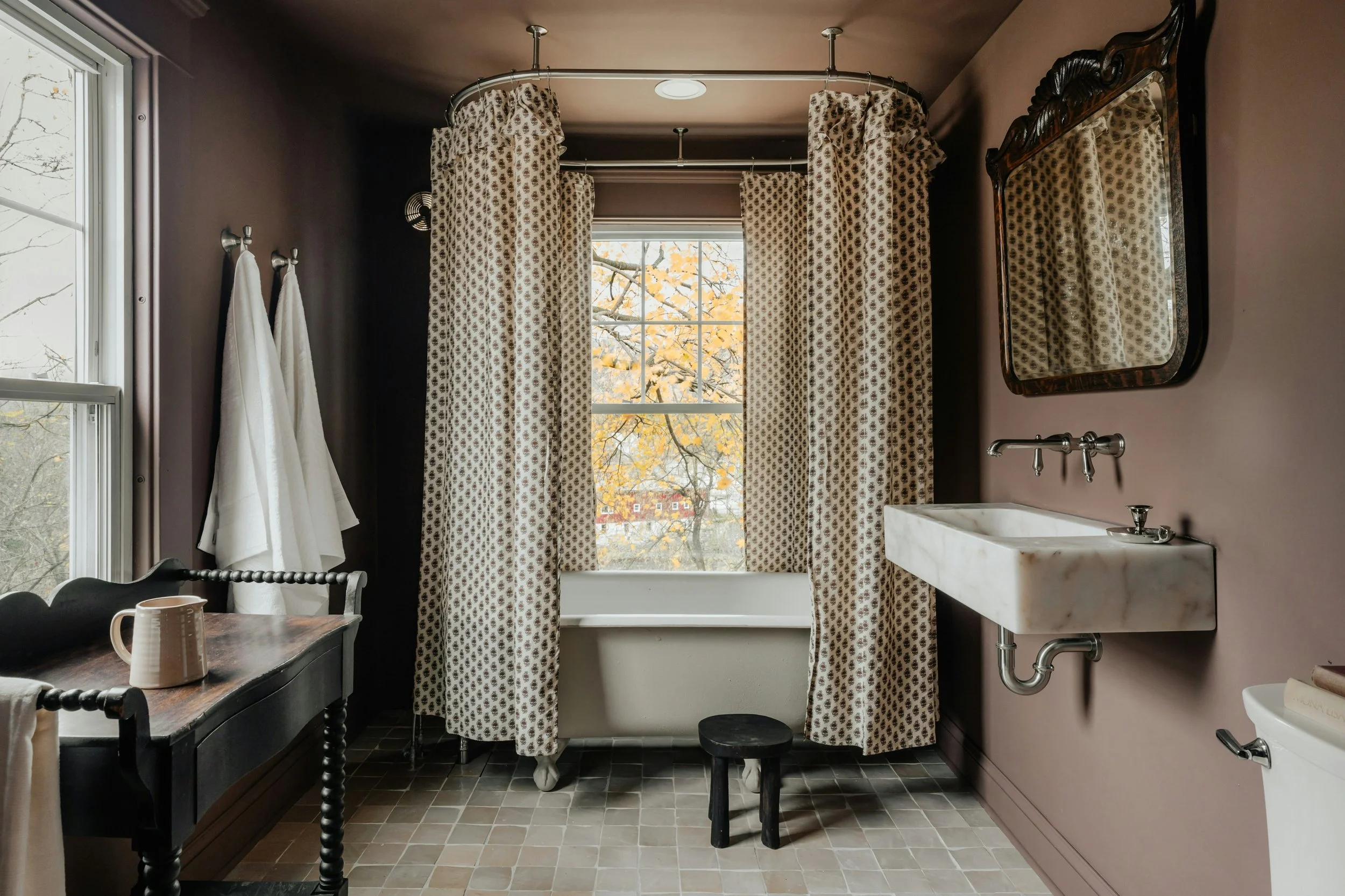 Bathroom with a bathtub behind patterned curtains, a window showing fall foliage, a wall-mounted sink, mirror, and hanging towels.