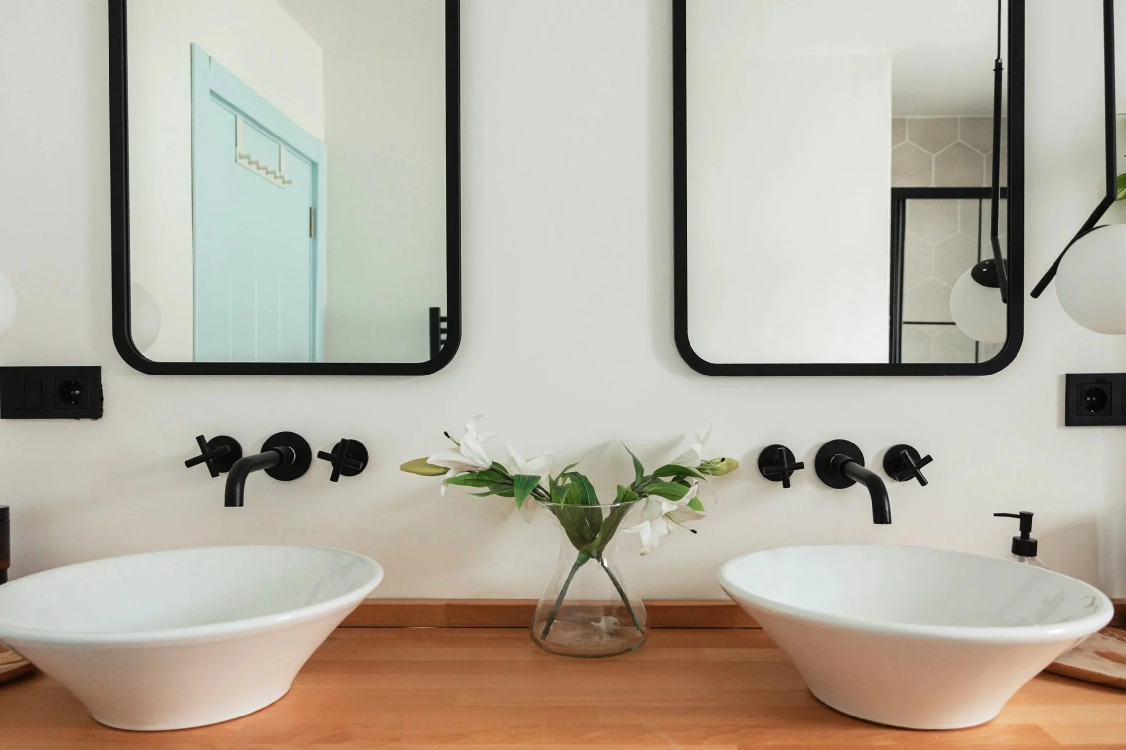 A bathroom vanity with two vessel sinks, black wall-mounted faucets, two rectangular mirrors with black frames, a clear glass vase with white lilies between the sinks, and a black soap dispenser. There are electrical outlets on each side of the mirrors, and a modern white light fixture with round globes is partially visible on the right side.