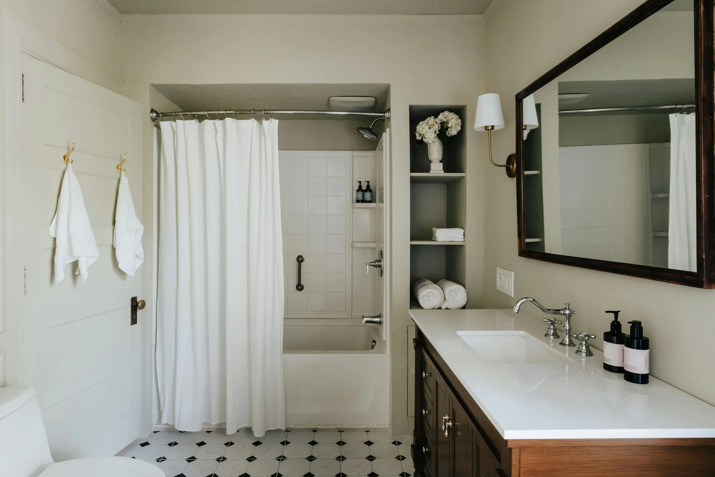 Bathroom with white walls, a bathtub with shower curtain, a vanity with a large mirror, and shelves with towels and flowers.