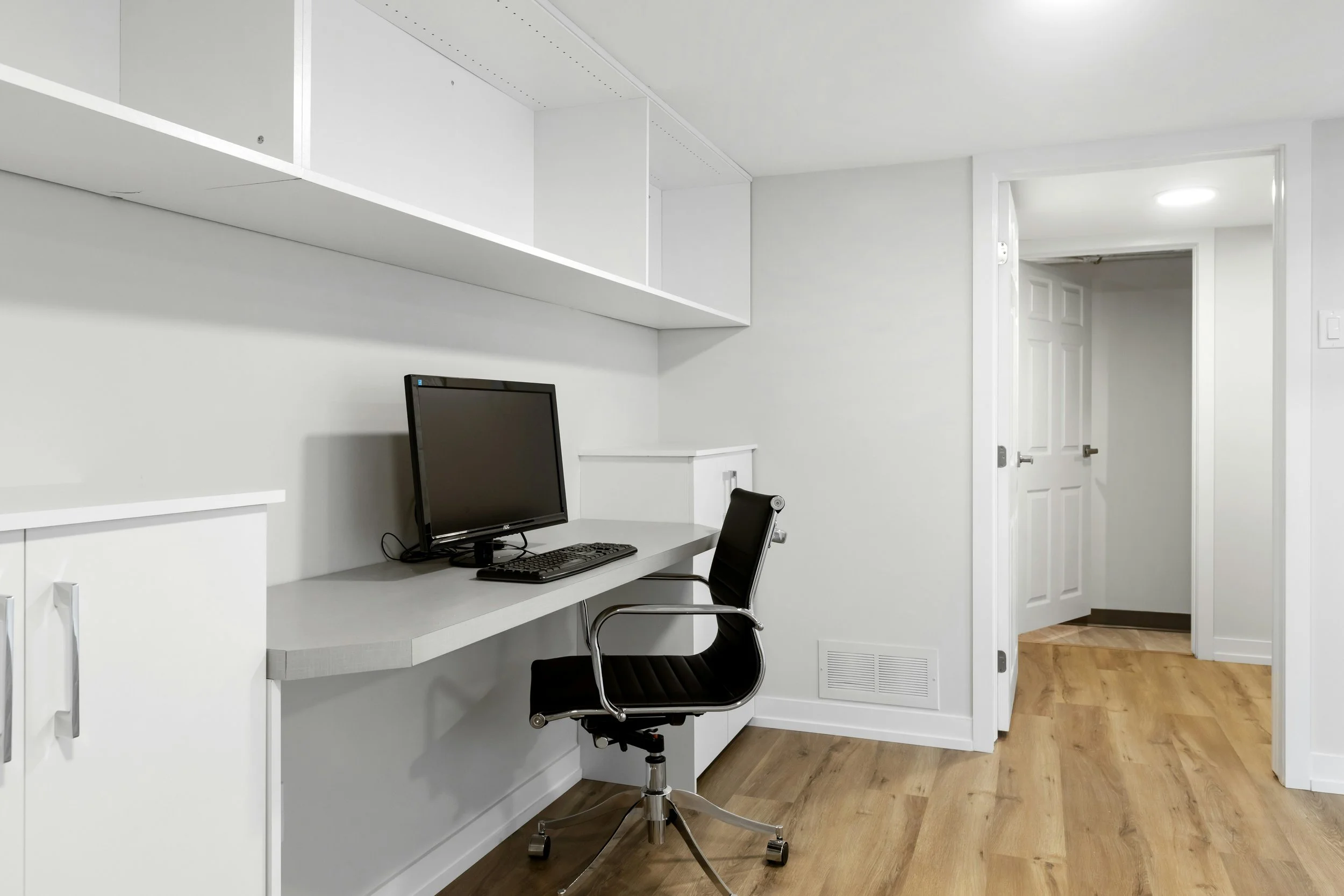 A minimalist home office with a black office chair, a desktop computer monitor, and a white built-in desk and shelves in a neutrally decorated room with wood flooring.