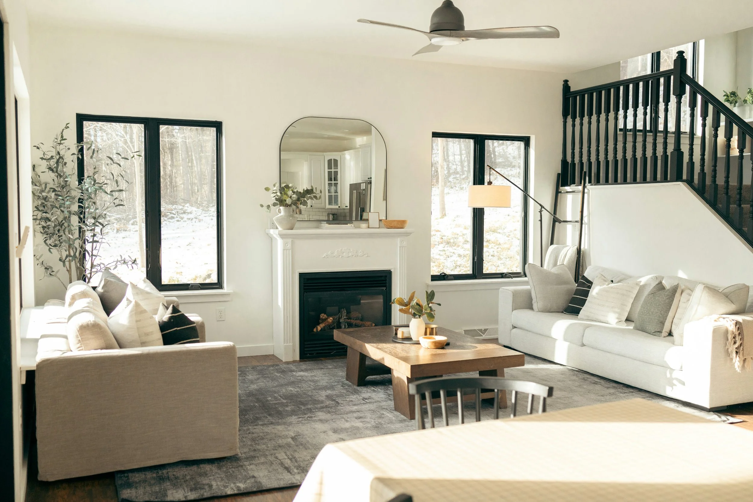 Bright living room with beige sofas, a wooden coffee table, a fireplace, and large windows showing a snowy landscape outside.