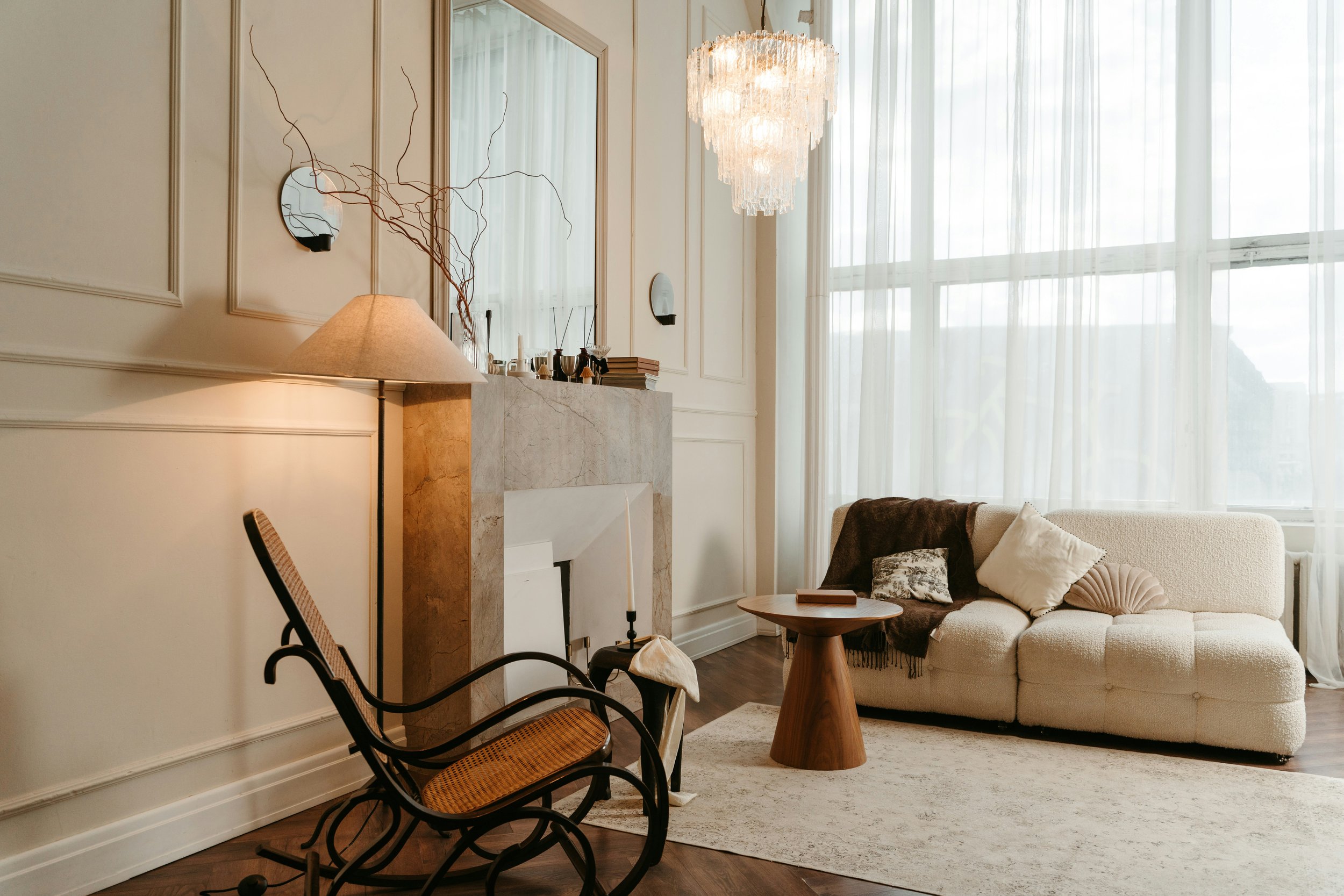 Living room with a beige sofa, dark brown blanket, and pillows, a small wooden side table, marble fireplace, large window with sheer curtains, a rocking chair, and a chandelier.