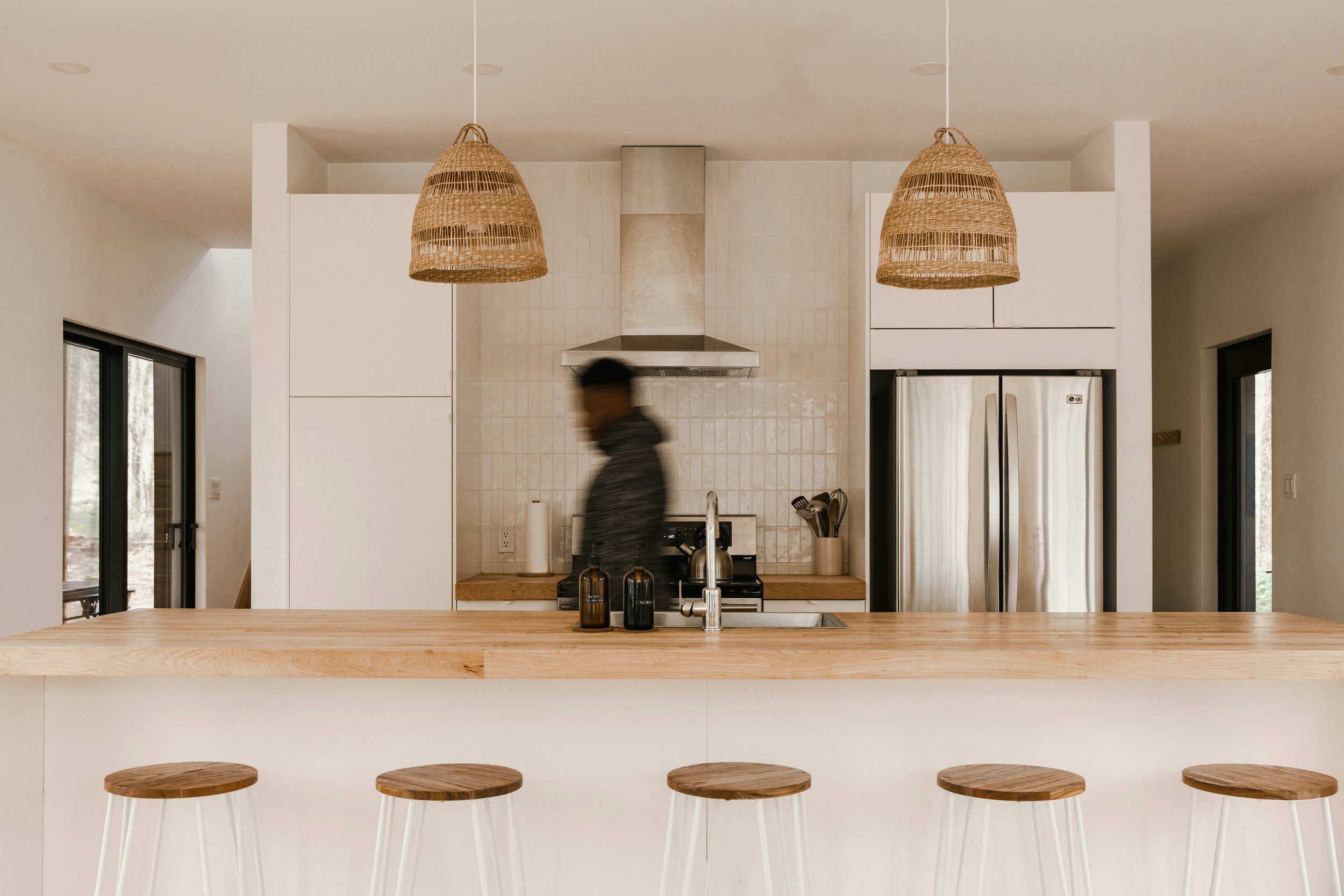 Modern kitchen with wooden countertops, stainless steel appliances, hanging wicker lights, and a blurred person walking across the room.