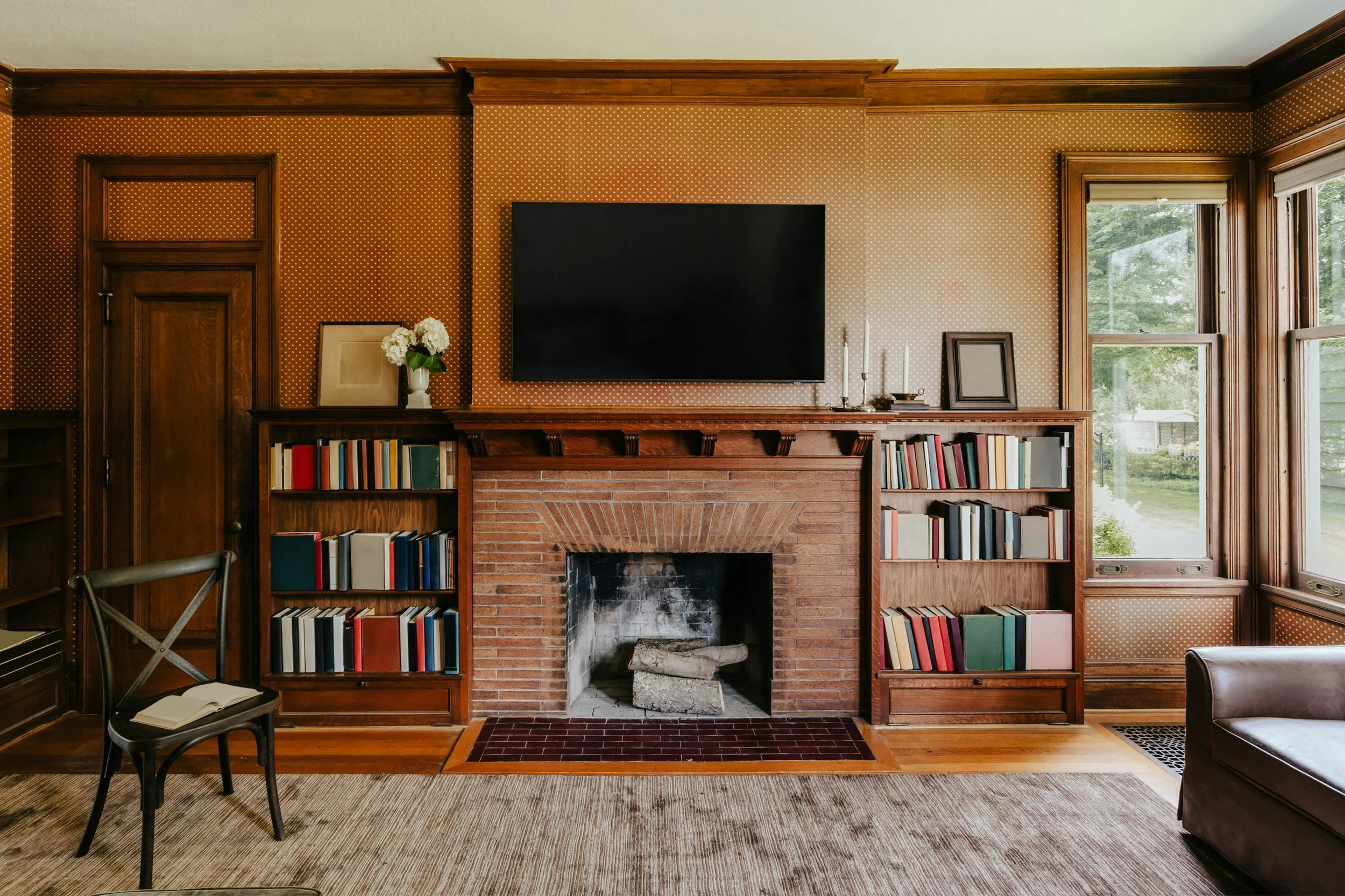 Living room with a brick fireplace, wooden bookshelves, and a mounted flat-screen TV. Large windows on the right let in natural light.